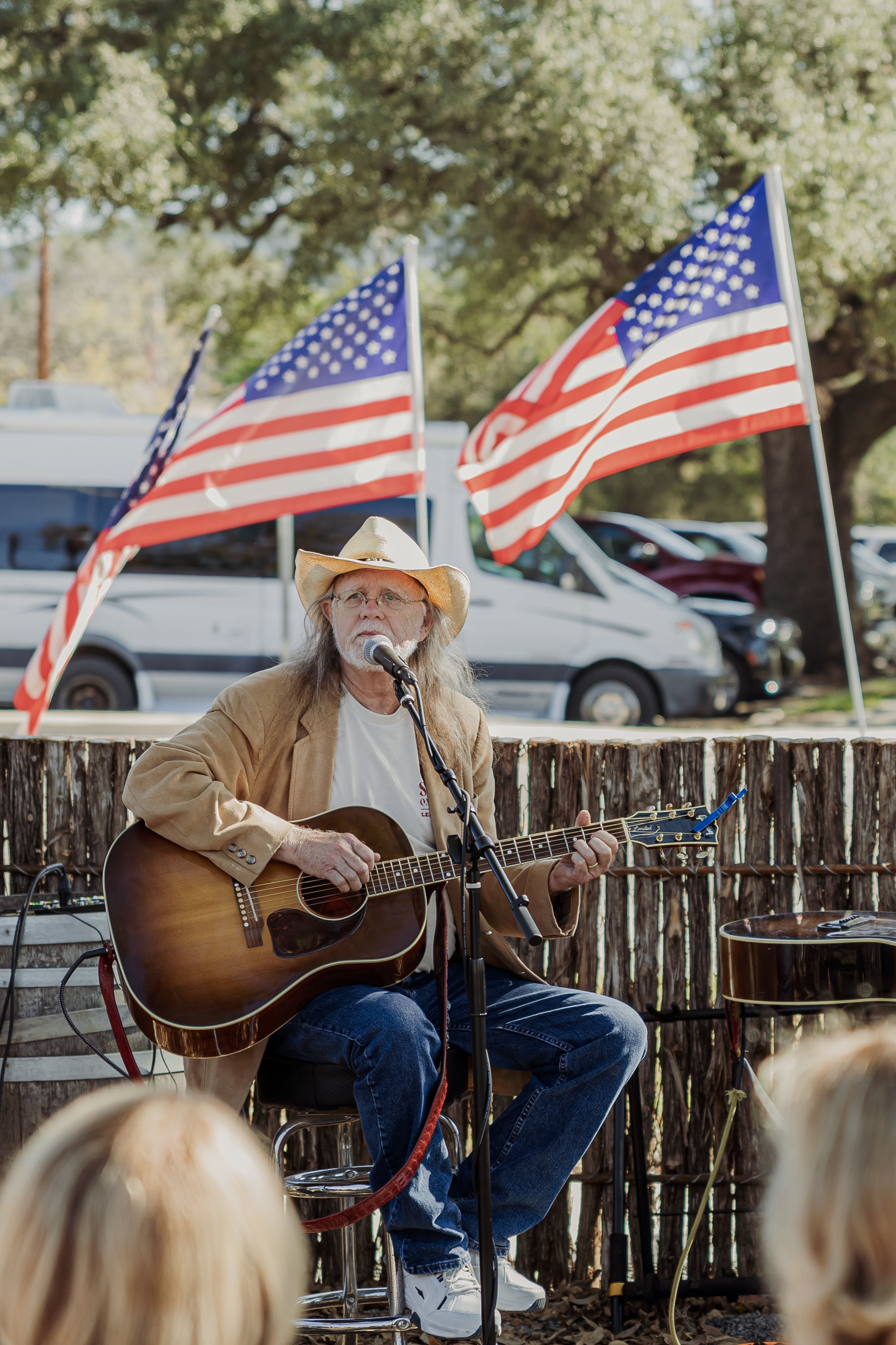 Man playing guitar and singing into microphone outdoors, with two American flags, parked cars, and trees in the background.