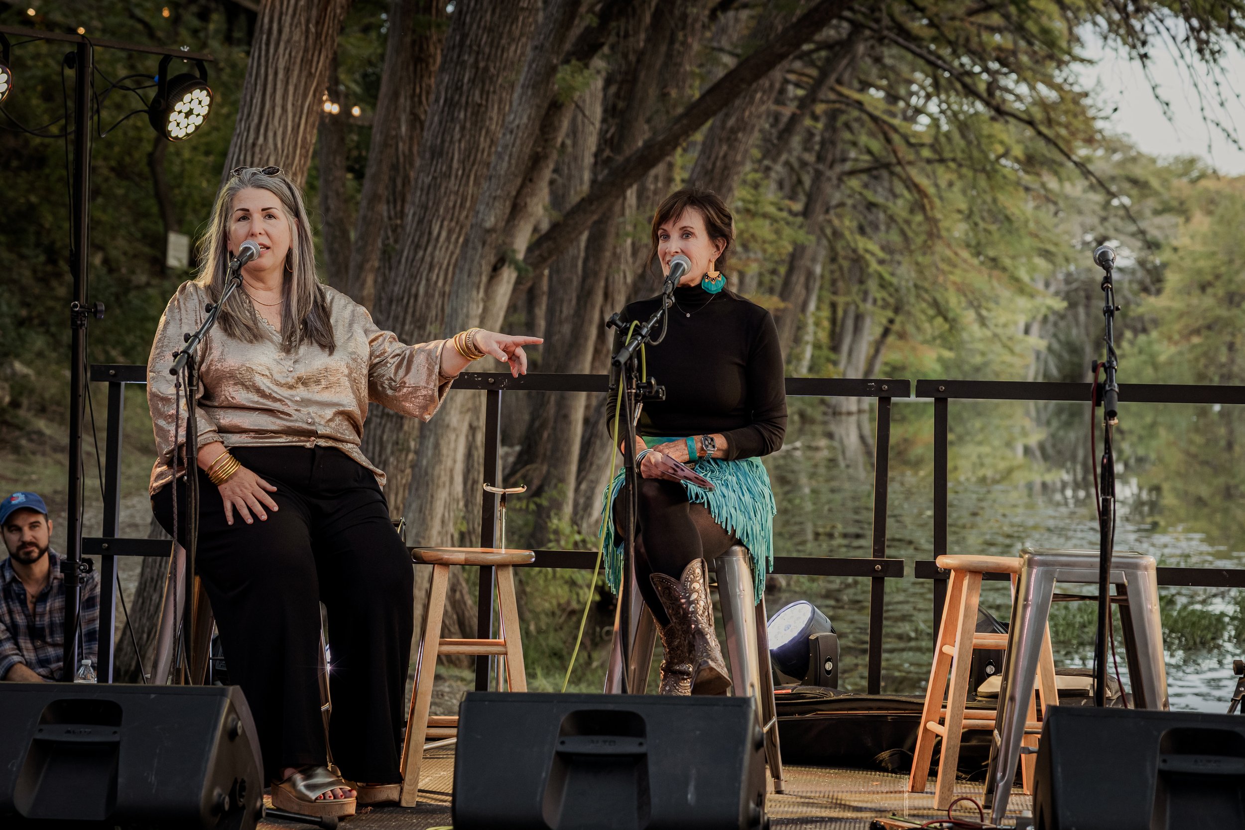 Two women speaking into microphones on an outdoor stage with a wooded background, one wearing a shiny beige top and black pants, the other in a black top and turquoise skirt, sitting on stools, with a man in a cap sitting in the background.
