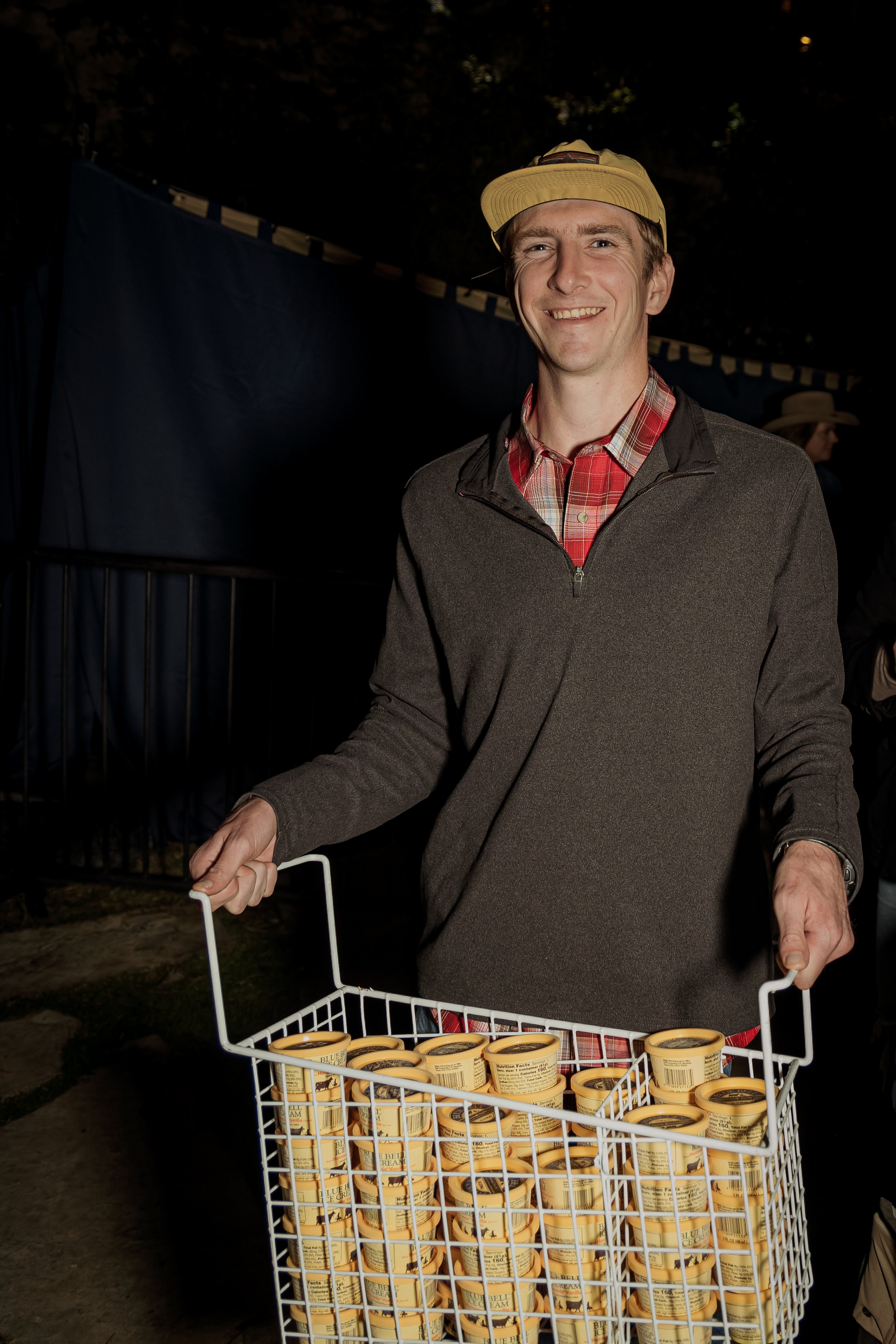 A man wearing a yellow cap and a gray jacket holding a white wire basket filled with small containers of ice cream at night.