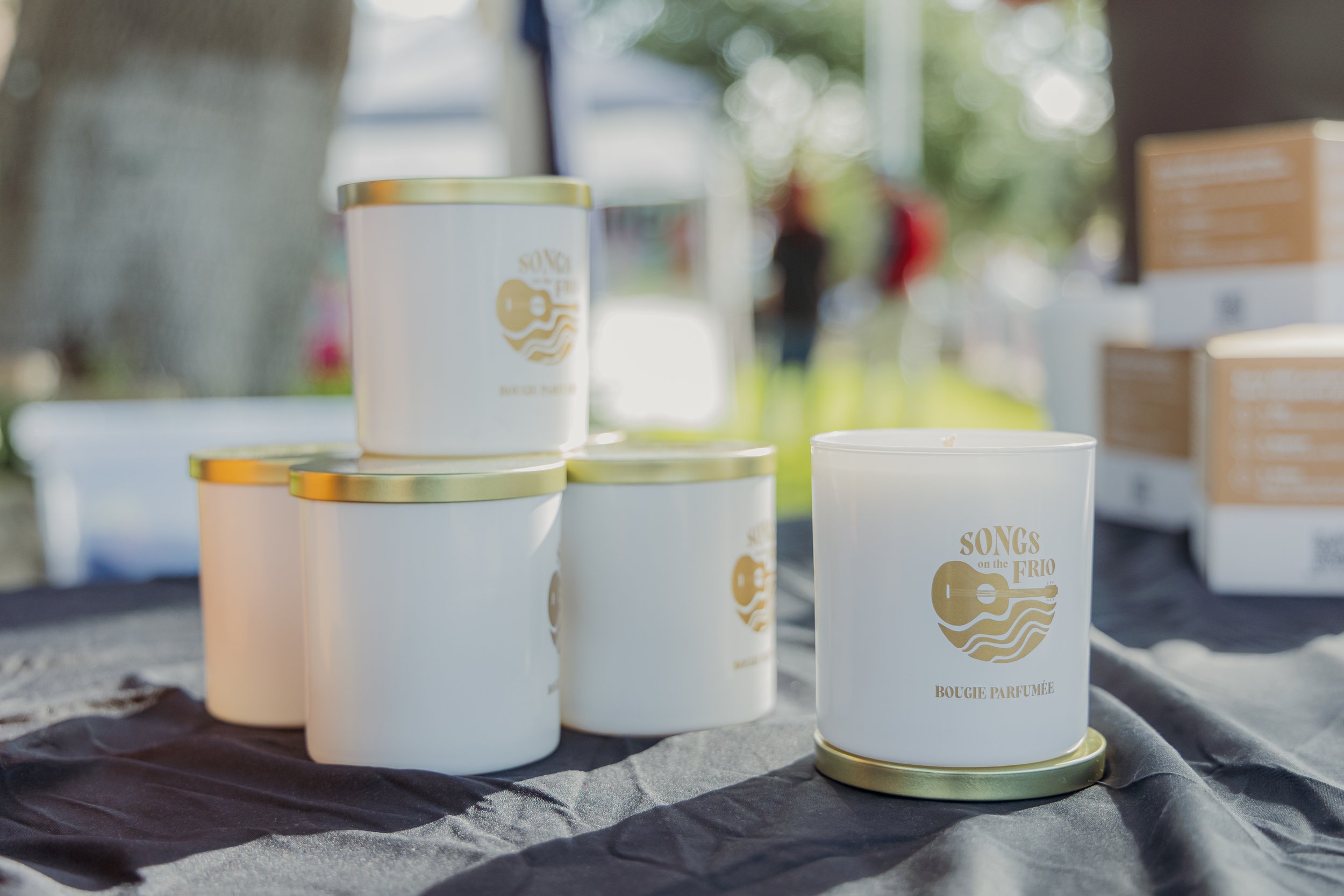 White scented candles with gold lids on a black cloth, featuring a gold guitar logo with waves and the words 'Songs on the Frio' and 'Bougie Parfumée' in gold lettering.