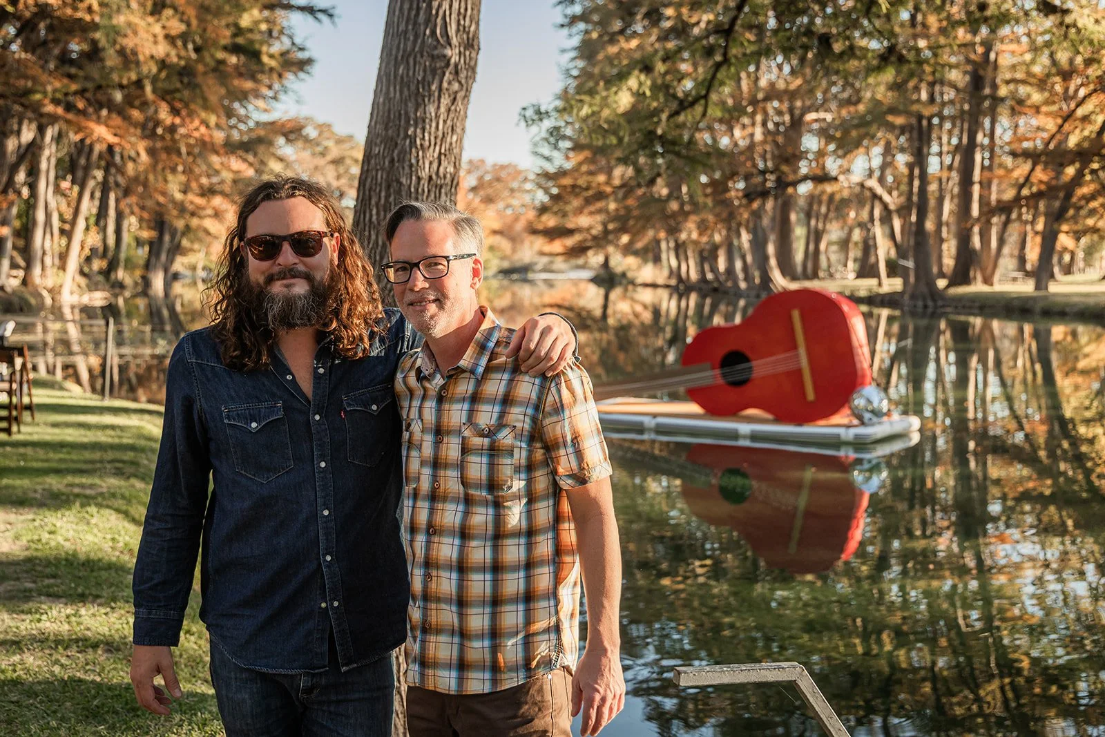 Two men standing by a lake surrounded by autumn trees, one with long curly hair and sunglasses, the other with short gray hair and glasses, both smiling. A red guitar-shaped paddleboard floats on the water in the background.