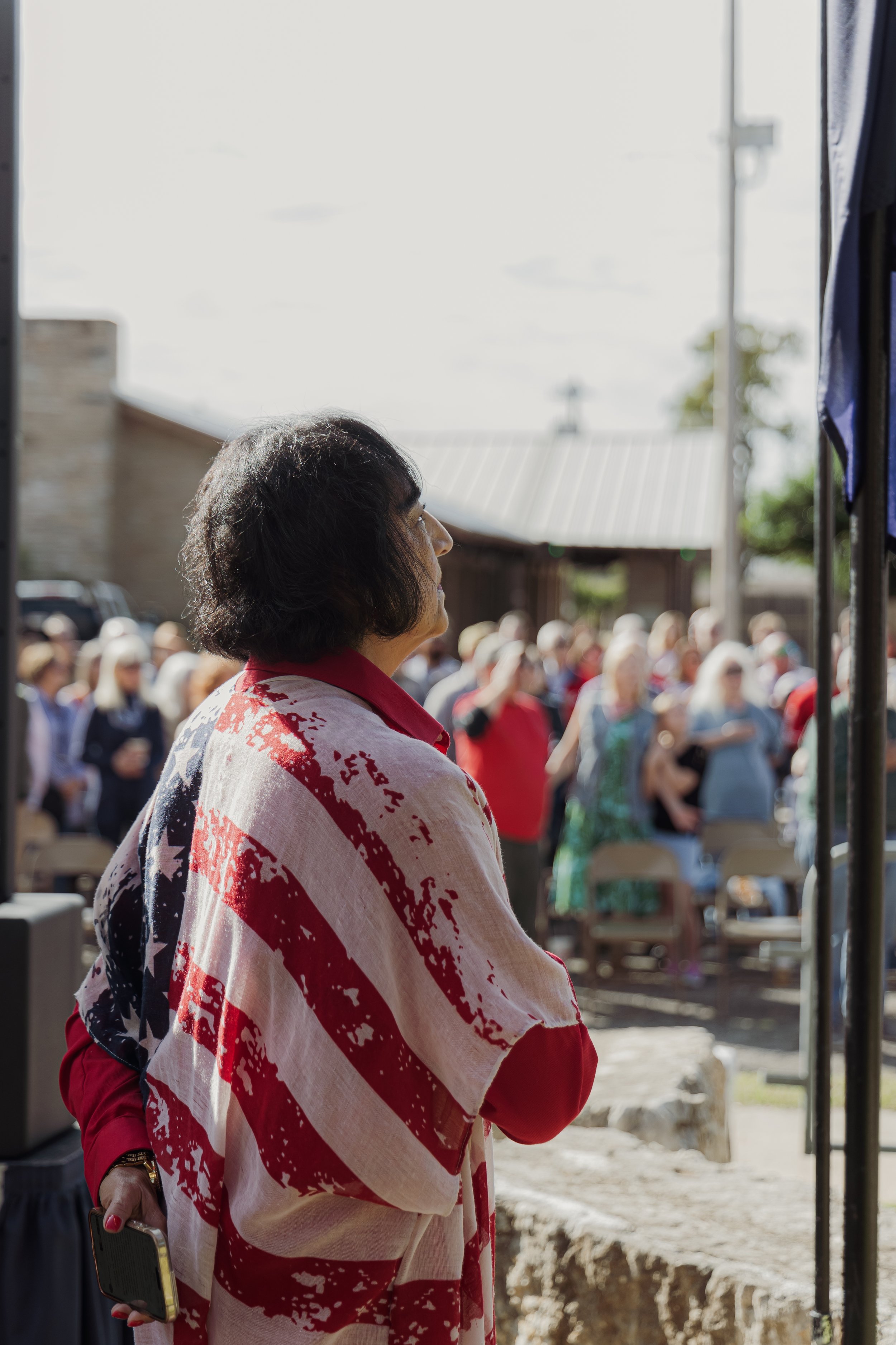 A woman standing outdoors, wearing a red, white, and blue patterned shawl, observing a crowd of people at an event.