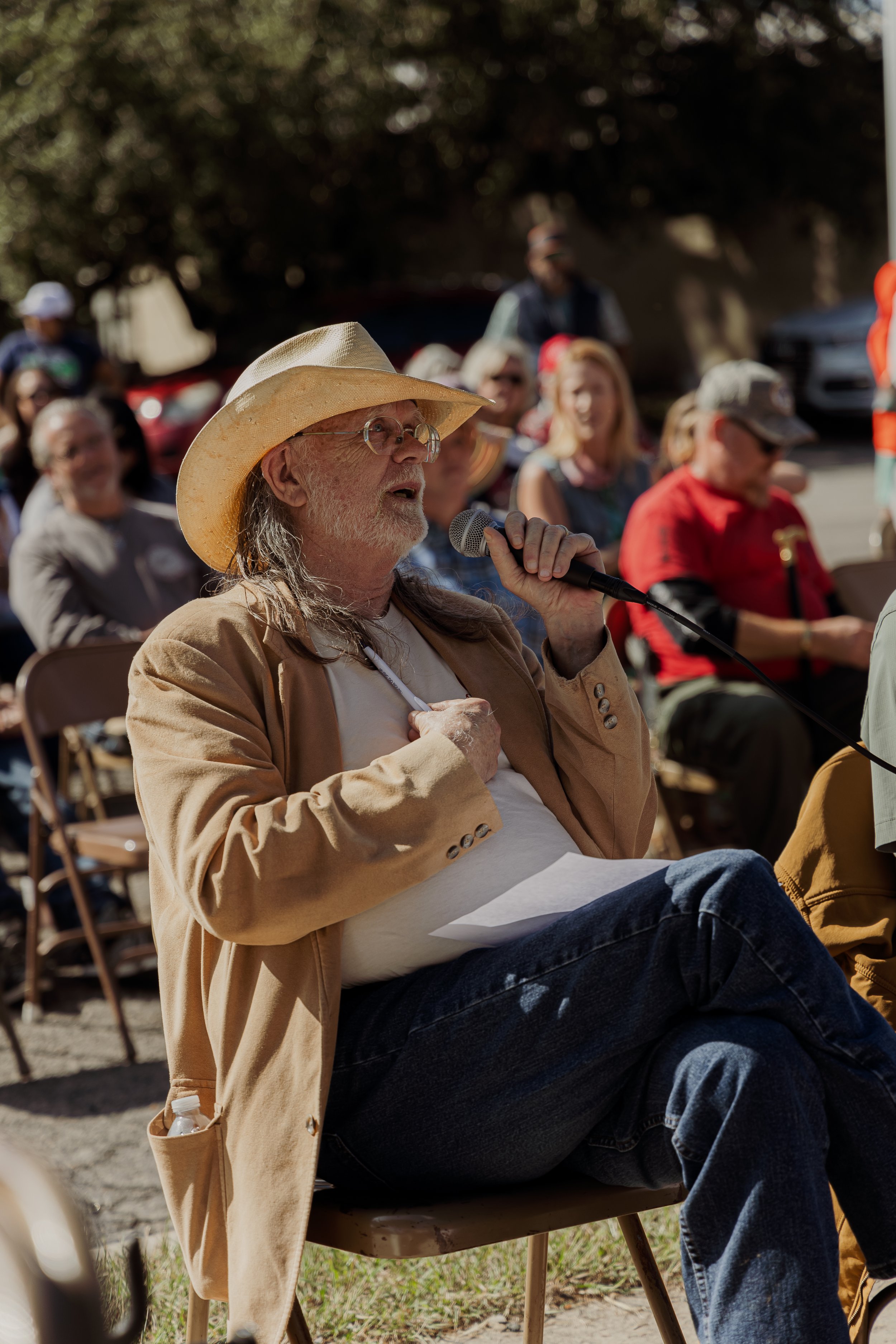 An elderly man wearing a cowboy hat and glasses, sitting outdoors among an audience, is speaking into a microphone during an event on a sunny day.