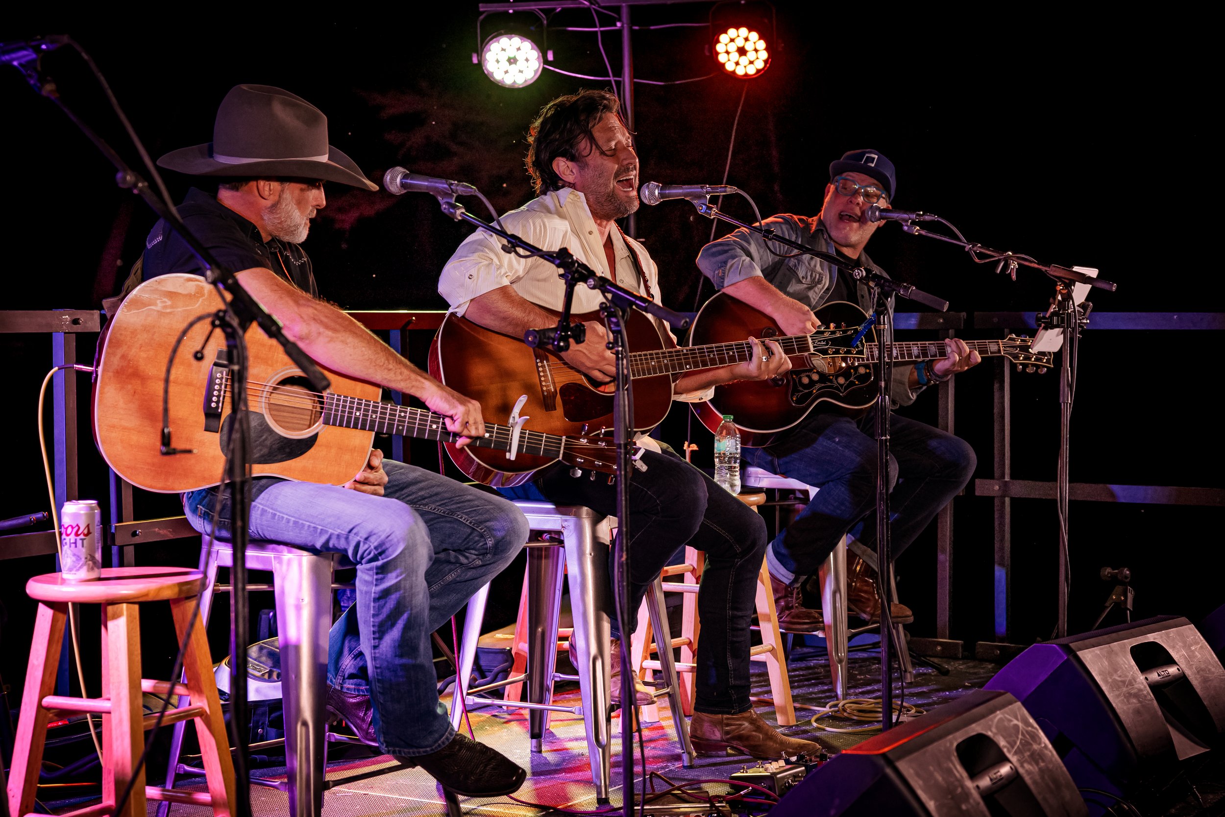 Three male musicians playing acoustic guitars on a stage with colorful lights, seated on stools, singing into microphones during a live performance.