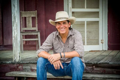 A woman wearing a cowboy hat and casual clothes sitting on the porch of a rustic building.