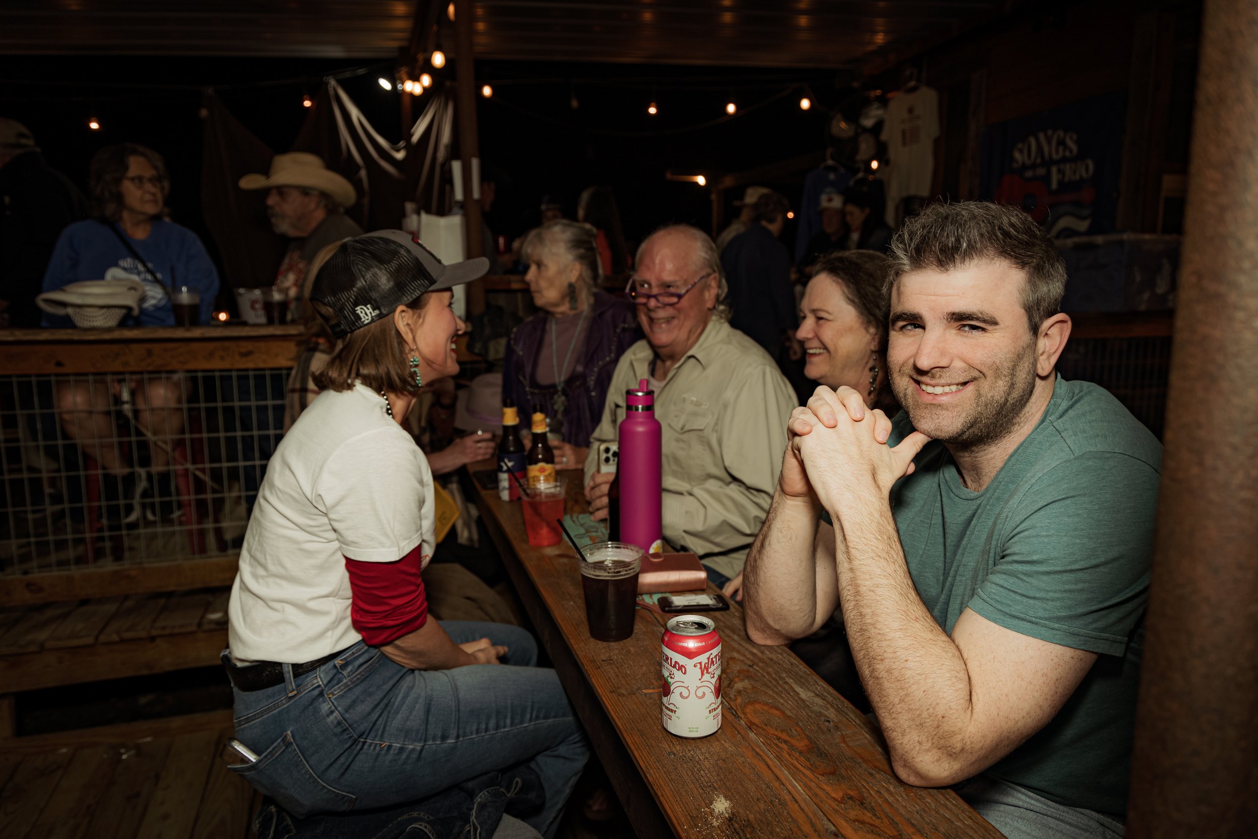 Group of people sitting at a wooden table in a lively, rustic bar or restaurant, smiling and enjoying conversation. There are drinks, notebooks, and a pink water bottle on the table.