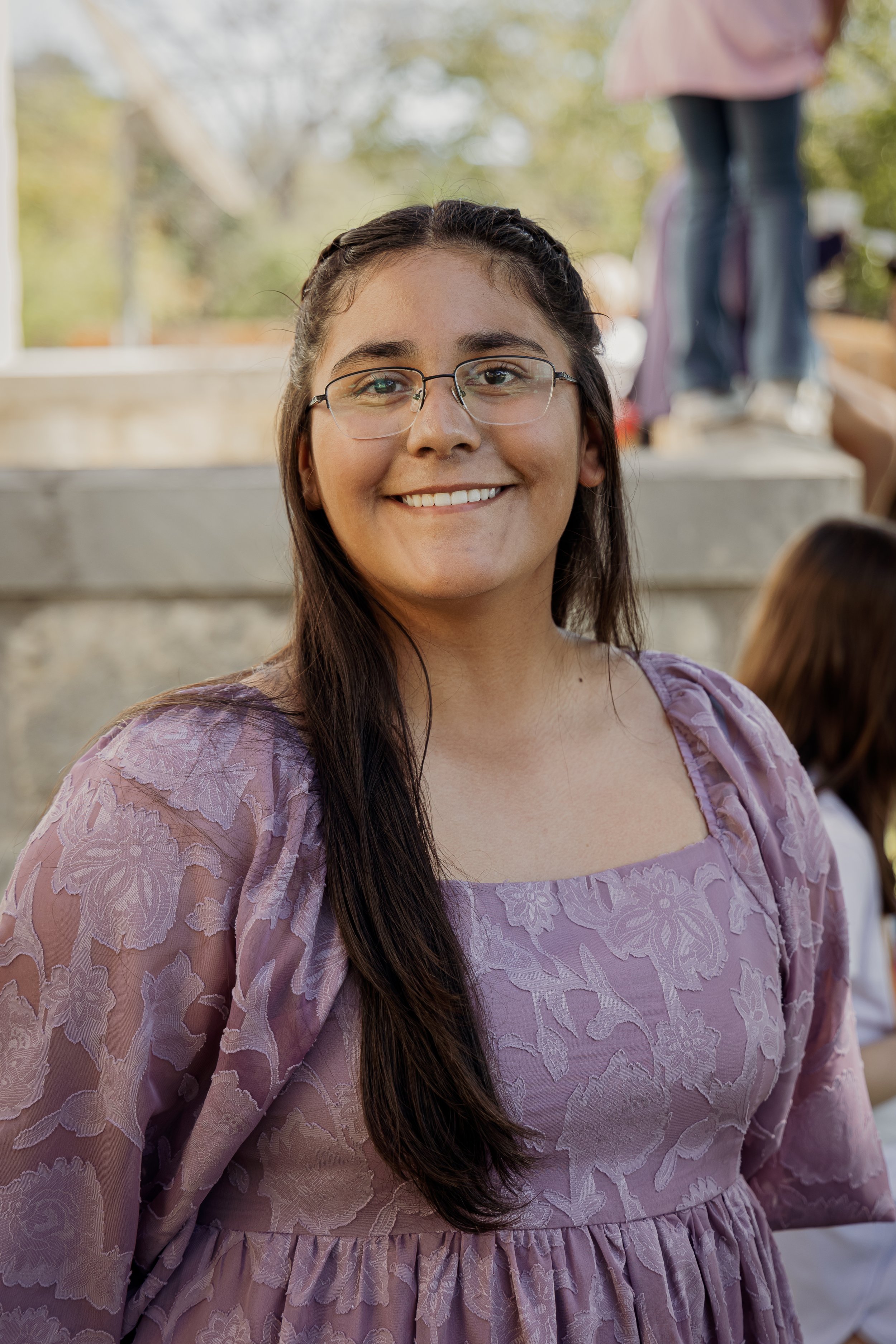 A young woman with long dark hair, glasses, and a lavender dress smiling outdoors.