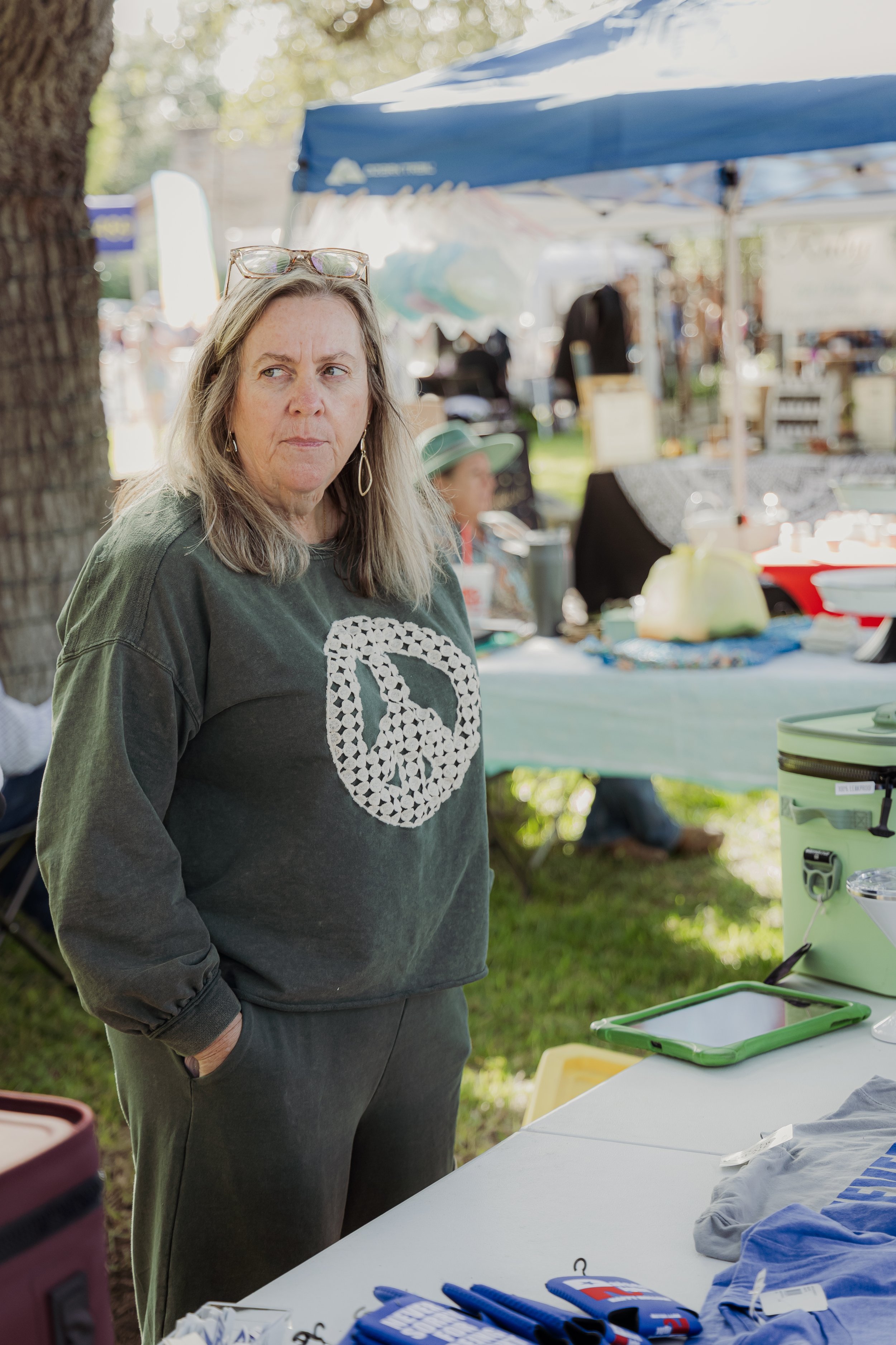 A woman with blonde hair, wearing a dark sweatshirt with a peace sign made of small white circles, standing outdoors at a flea market or craft fair under a large tree with tents and tables in the background.