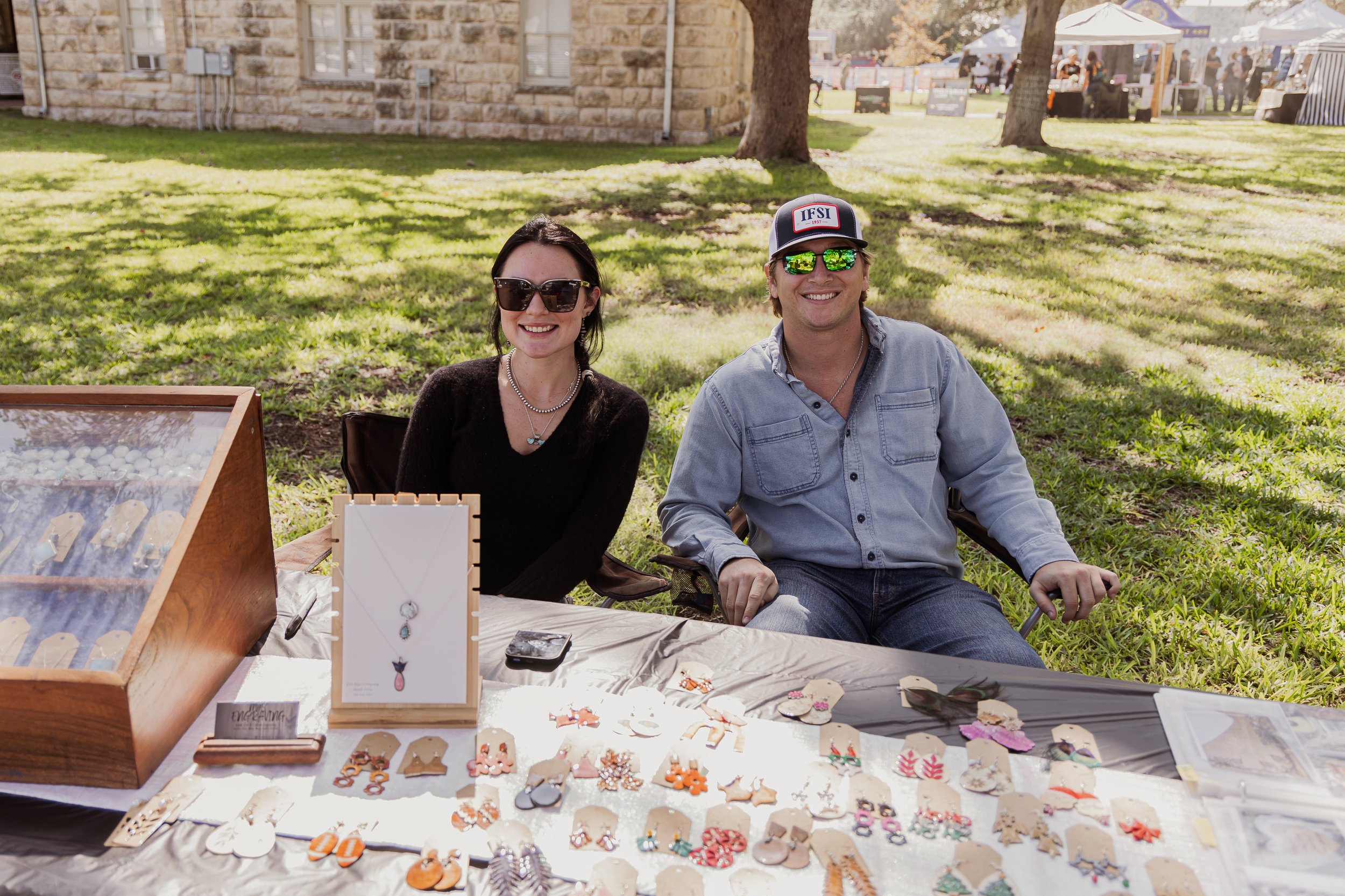 Two smiling people sitting at a jewelry booth outdoors with various necklaces, earrings, and jewelry items on display.
