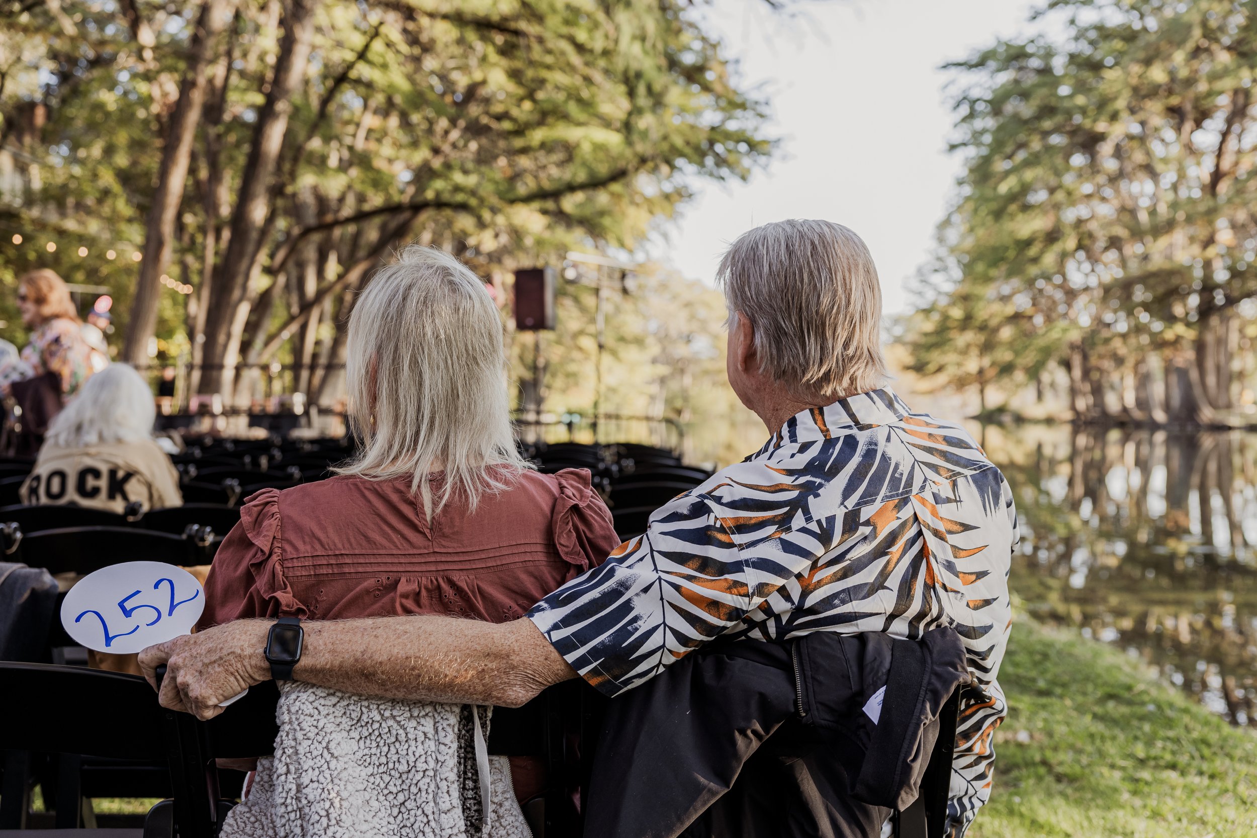 Older man and woman sitting together outdoors near a body of water, with trees and string lights in the background, embracing and watching a performance.