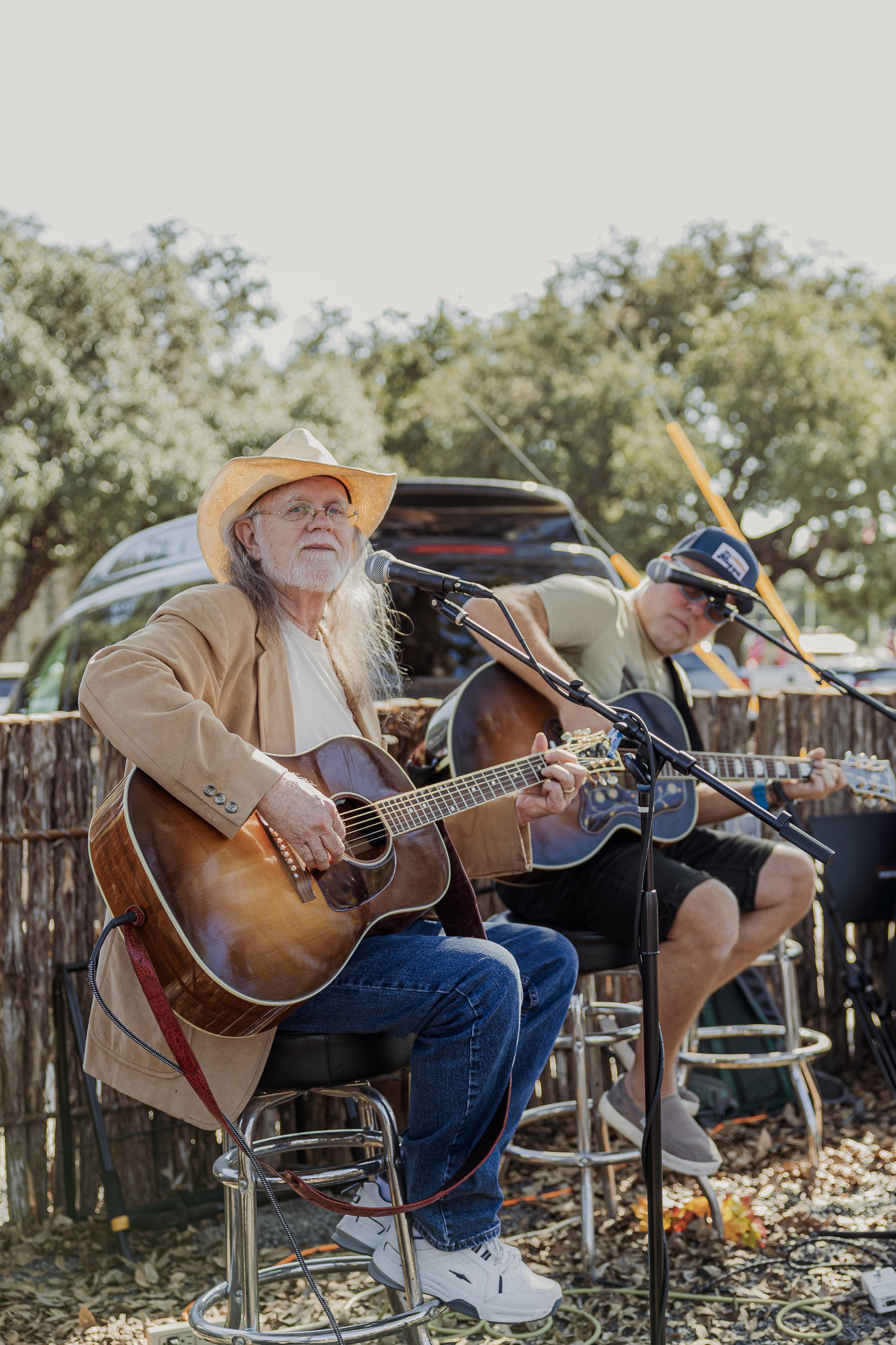 Two men playing acoustic guitars outdoors during a music performance, with one older man wearing a cowboy hat and sunglasses and the other man wearing a baseball cap and sunglasses, in front of a vehicle and surrounded by trees.