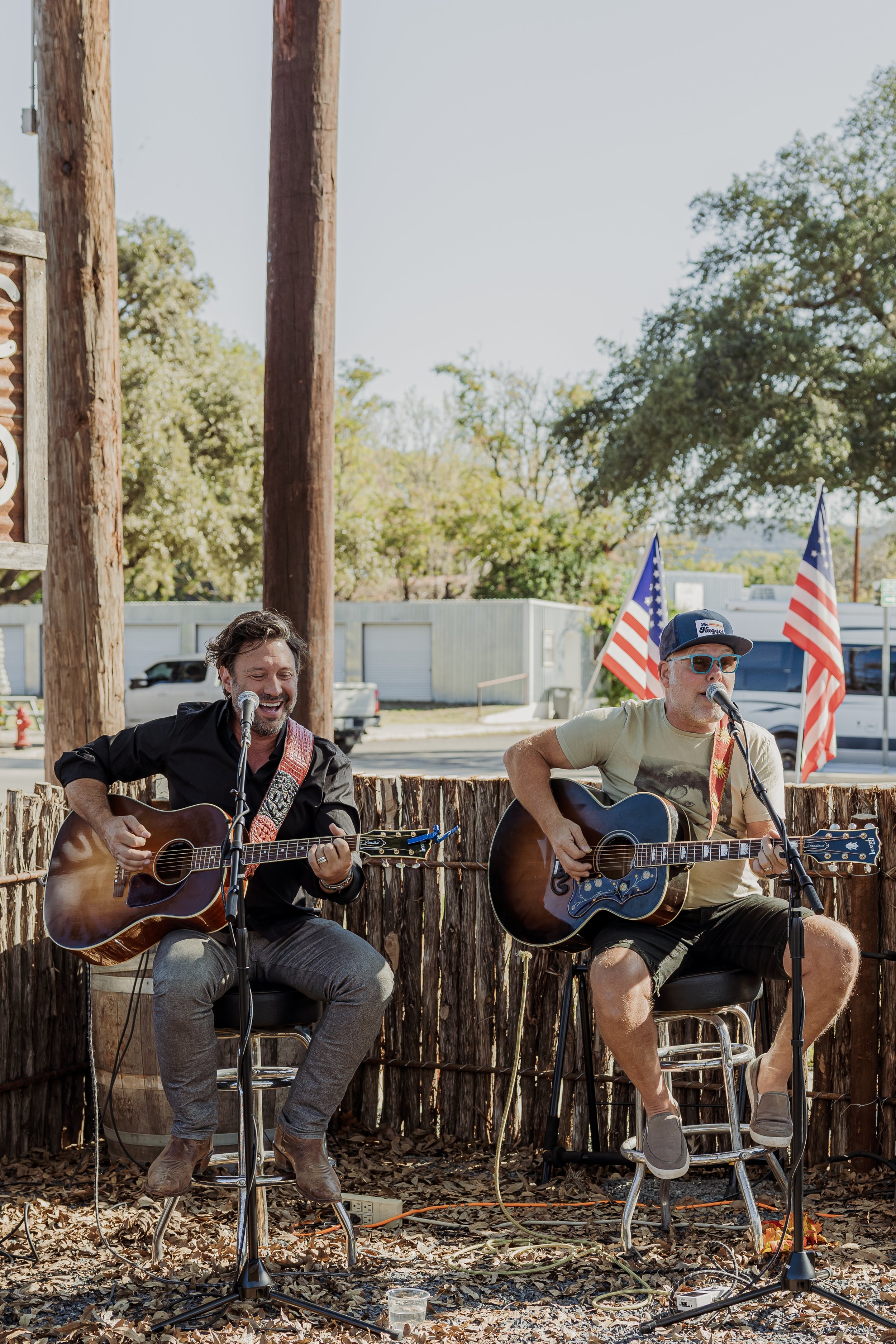Two men playing acoustic guitars and singing into microphones outdoors, with American flags and trees in the background.