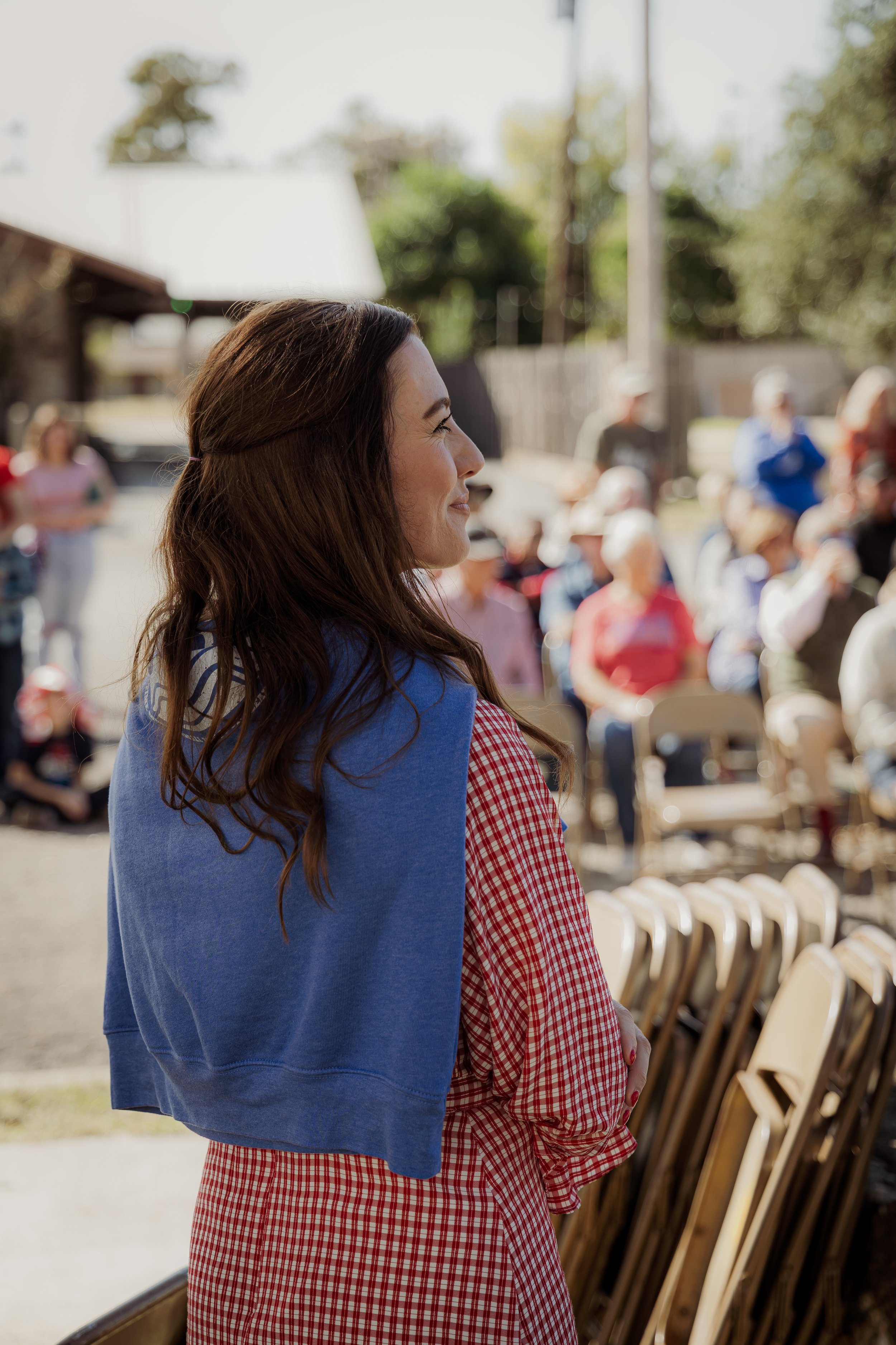 A woman with long brown hair, wearing a red checkered dress and a blue sweatshirt draped over her shoulders, stands outdoors in front of a seated crowd, smiling and looking to the side during daytime.