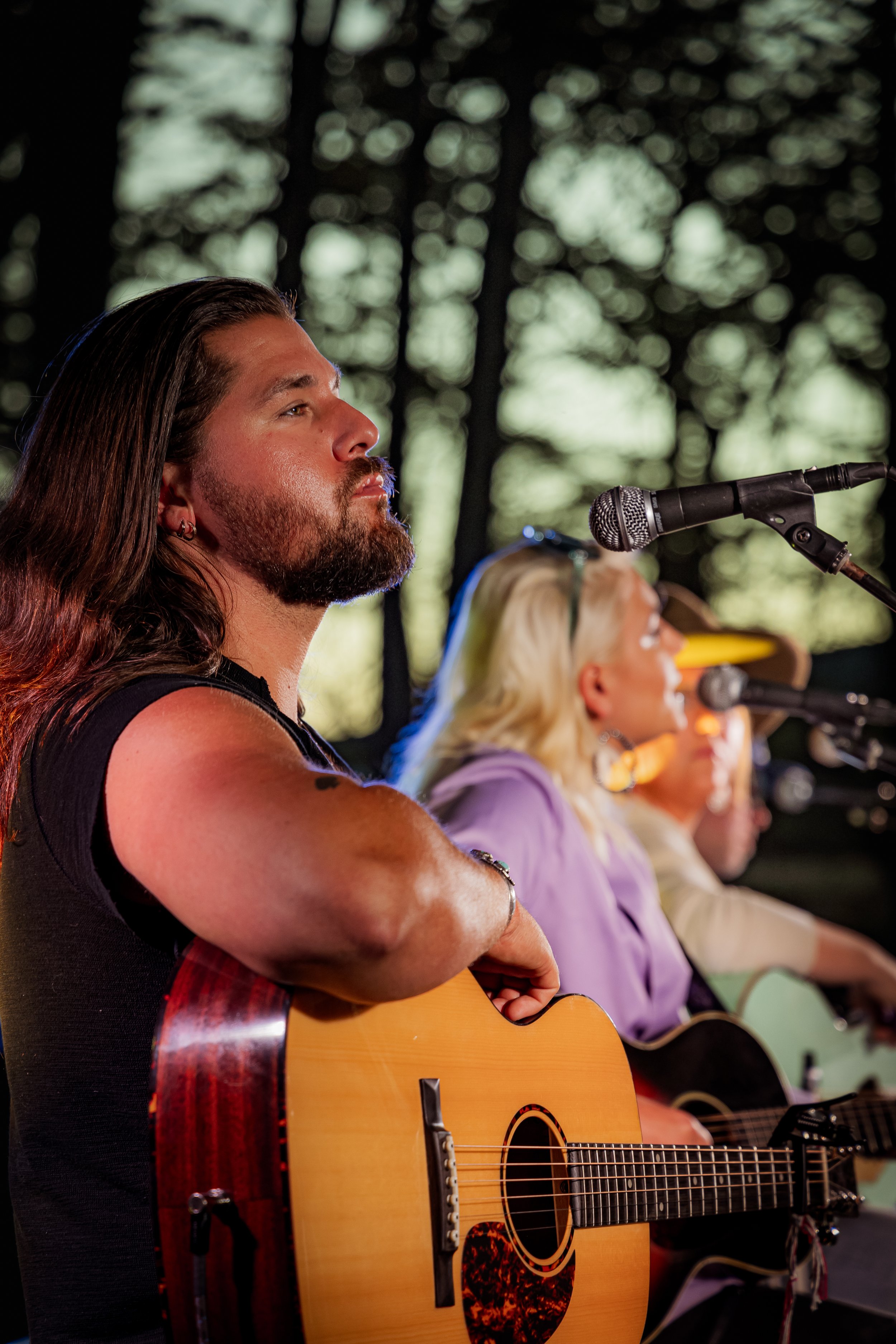 Musicians playing guitars and singing into microphones on stage outdoors at sunset.