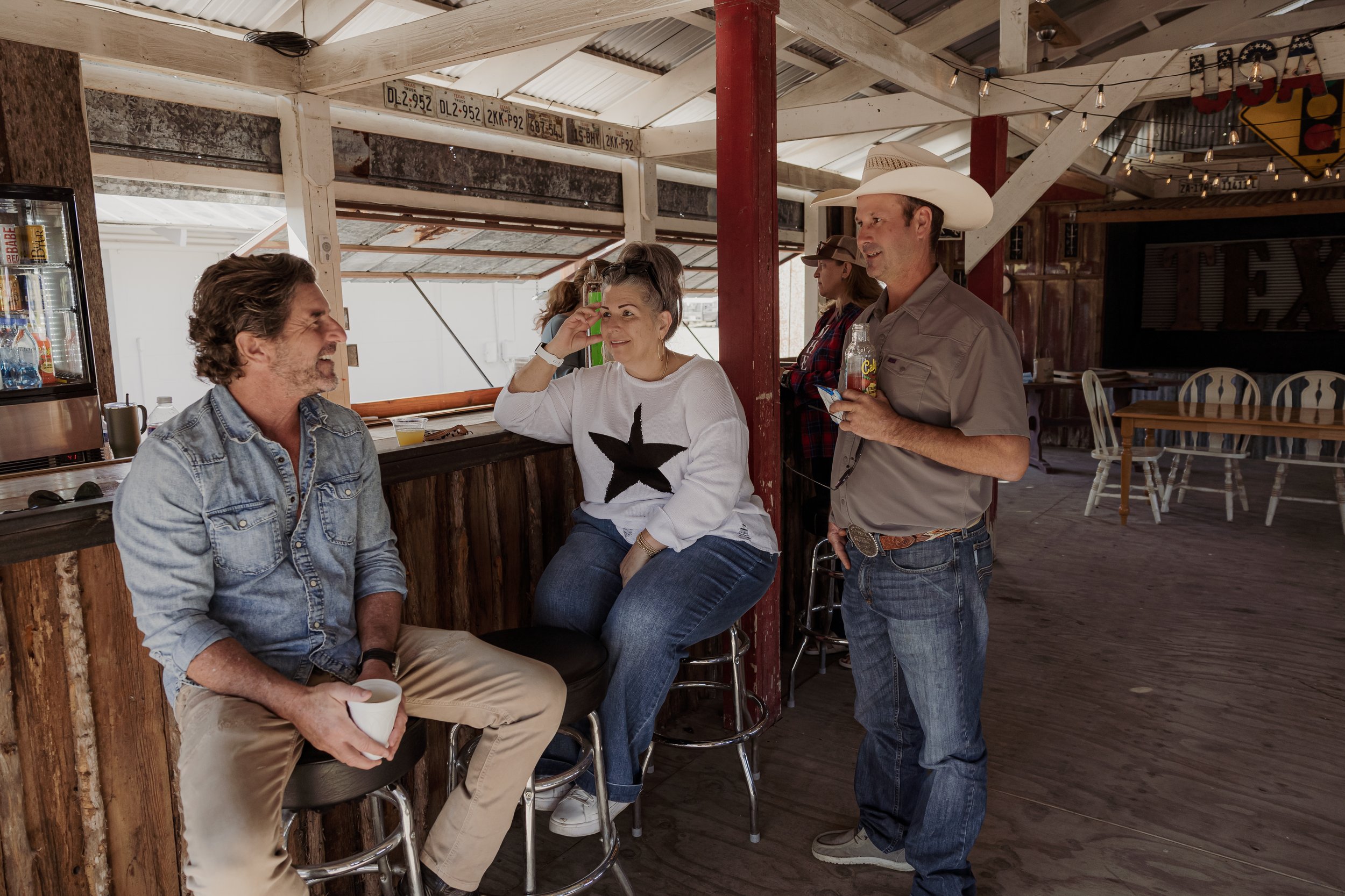 Four people socializing inside a rustic bar with wooden walls and flooring. A man in a denim jacket is sitting on a barstool, holding a cup, and talking to a woman in a white sweater with a star design, who is sitting on the counter. A man in a cowbo