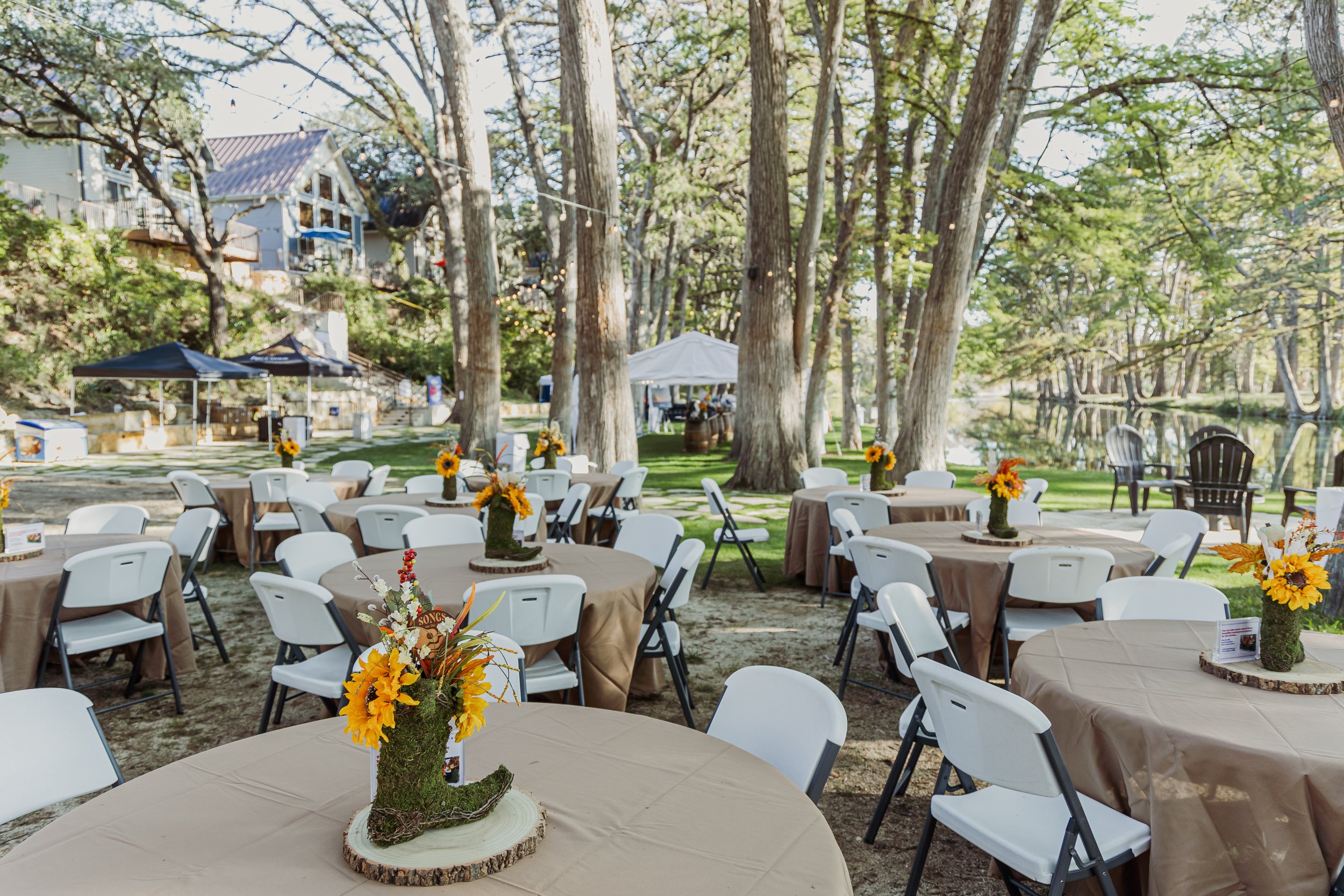 Outdoor event setup with round tables covered in tan tablecloths, decorated with sunflower centerpieces on wooden bases, set among trees near a lake or pond, with chairs arranged around the tables.