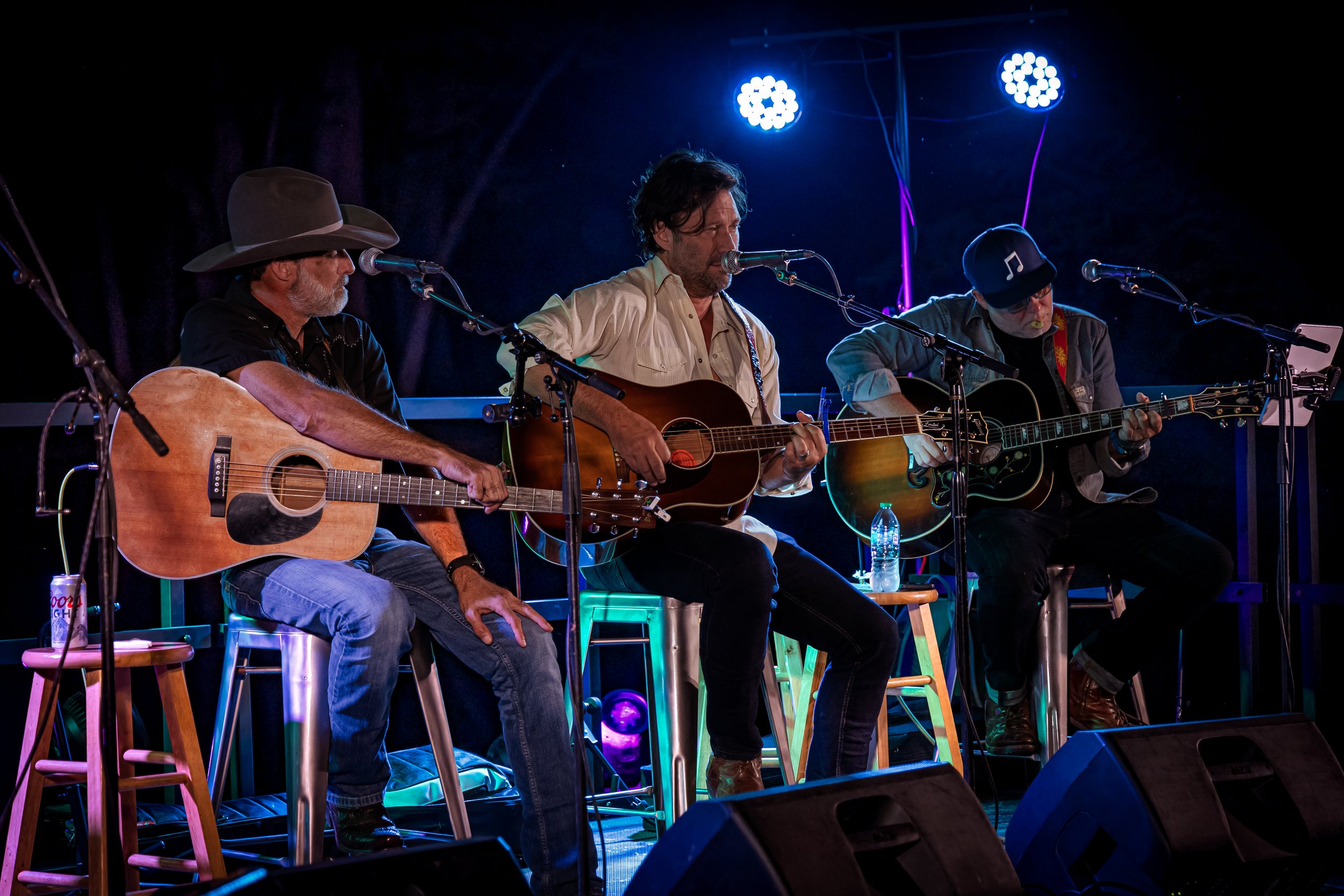 Three male musicians playing acoustic guitars on stage with microphones, illuminated by stage lights, with water bottles and stools nearby.