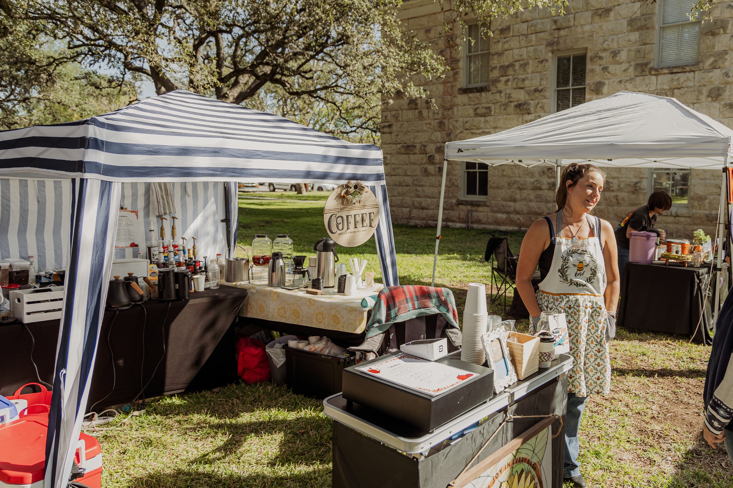 A woman standing outdoors under a white canopy at a coffee stand with various coffee-making equipment, a signage reading 'COFFEE,' and a second vendor behind a black table with drinks, with a large stone building in the background.