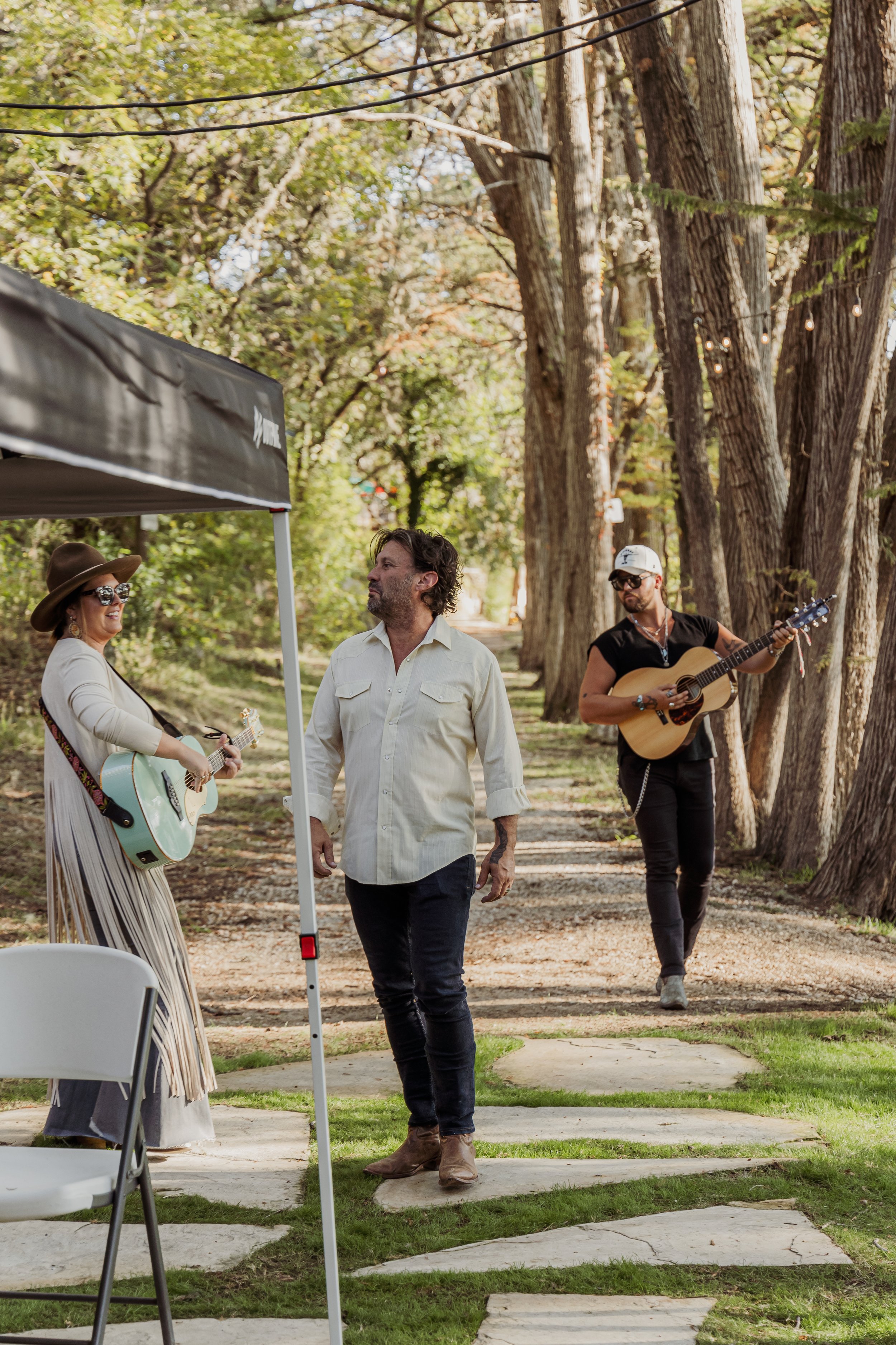 A musician smiling and playing an acoustic guitar, a man walking on a leaf-covered path, and another musician playing an acoustic guitar, all outside among tall trees with string lights overhead.