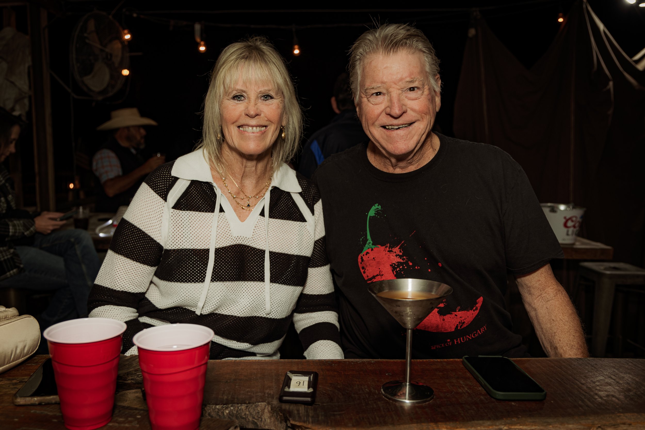 Smiling elderly woman and man sitting at a table with red cups, a smartphone, and a cocktail glass, in a dimly lit social setting.