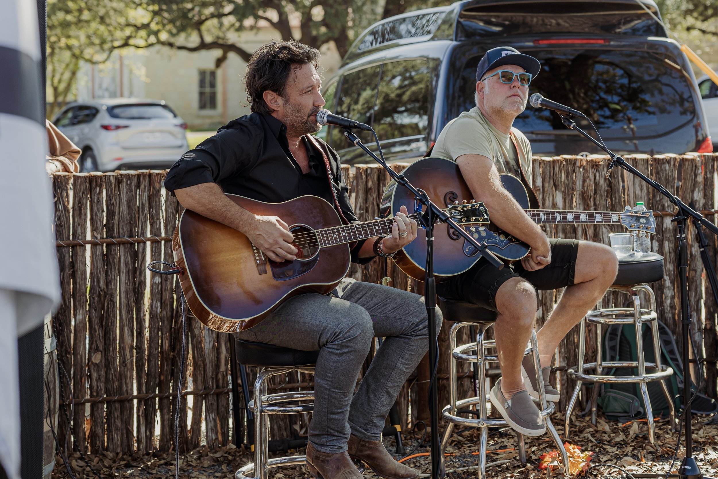 Two men playing acoustic guitars and singing outdoors, sitting on bar stools in front of a rustic wooden fence, with parked cars and trees in the background.