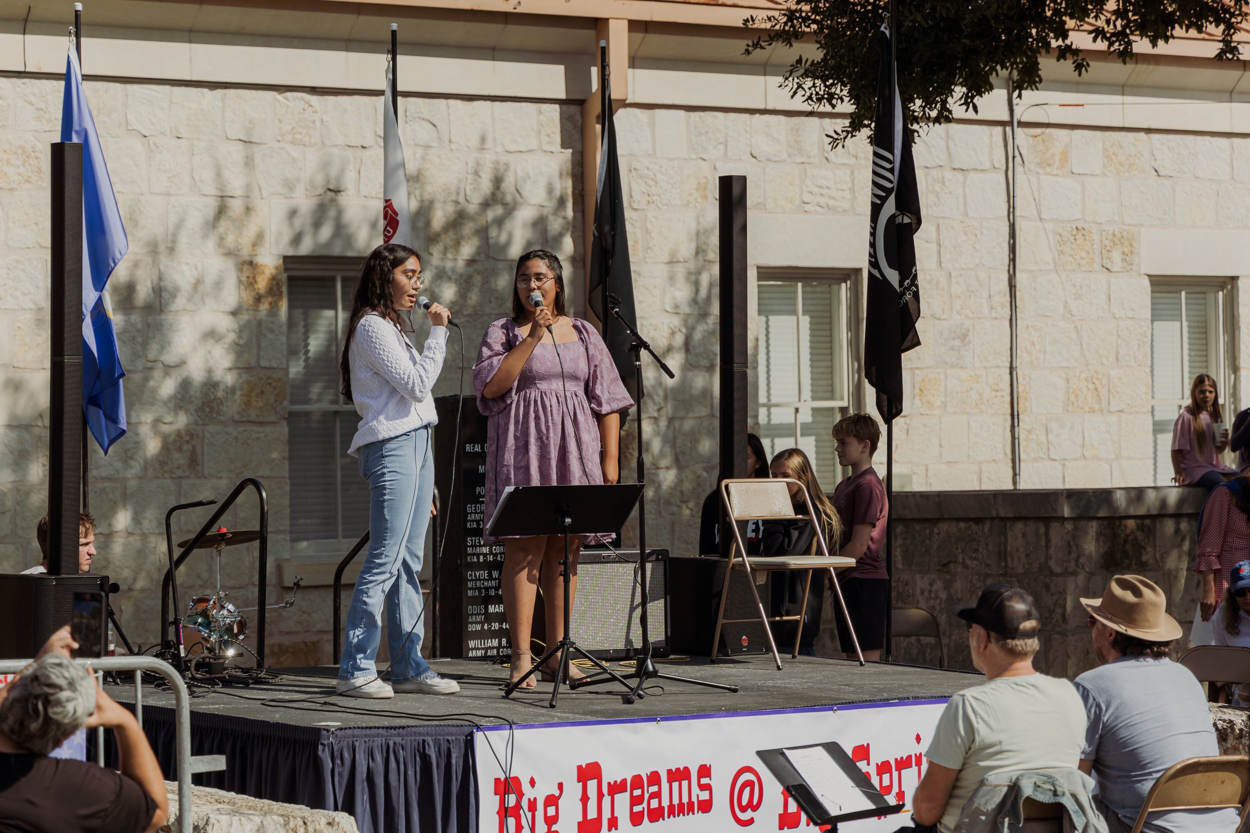 Two women stand on an outdoor stage singing into microphones. The stage has a banner with the words 'Rie Dreams @' partially visible. There are flags behind them and a group of people seated and standing nearby, watching the performance.