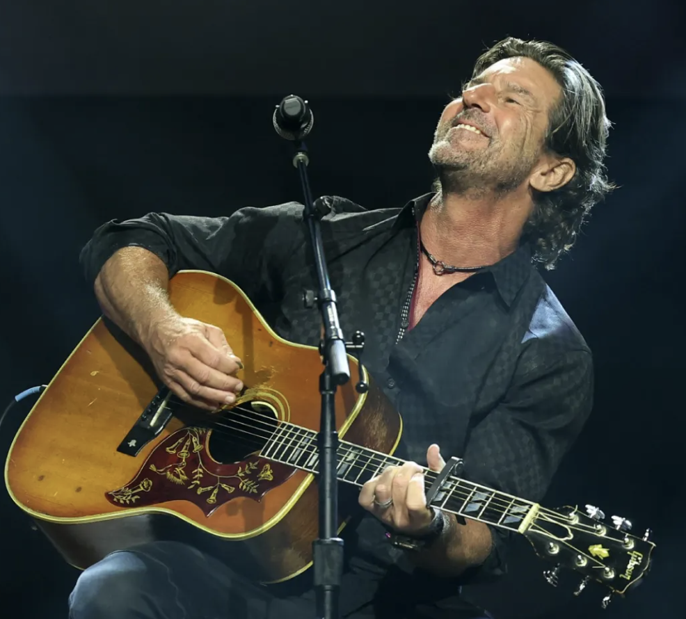 A man with gray hair and a beard playing an acoustic guitar on stage, smiling with eyes closed.