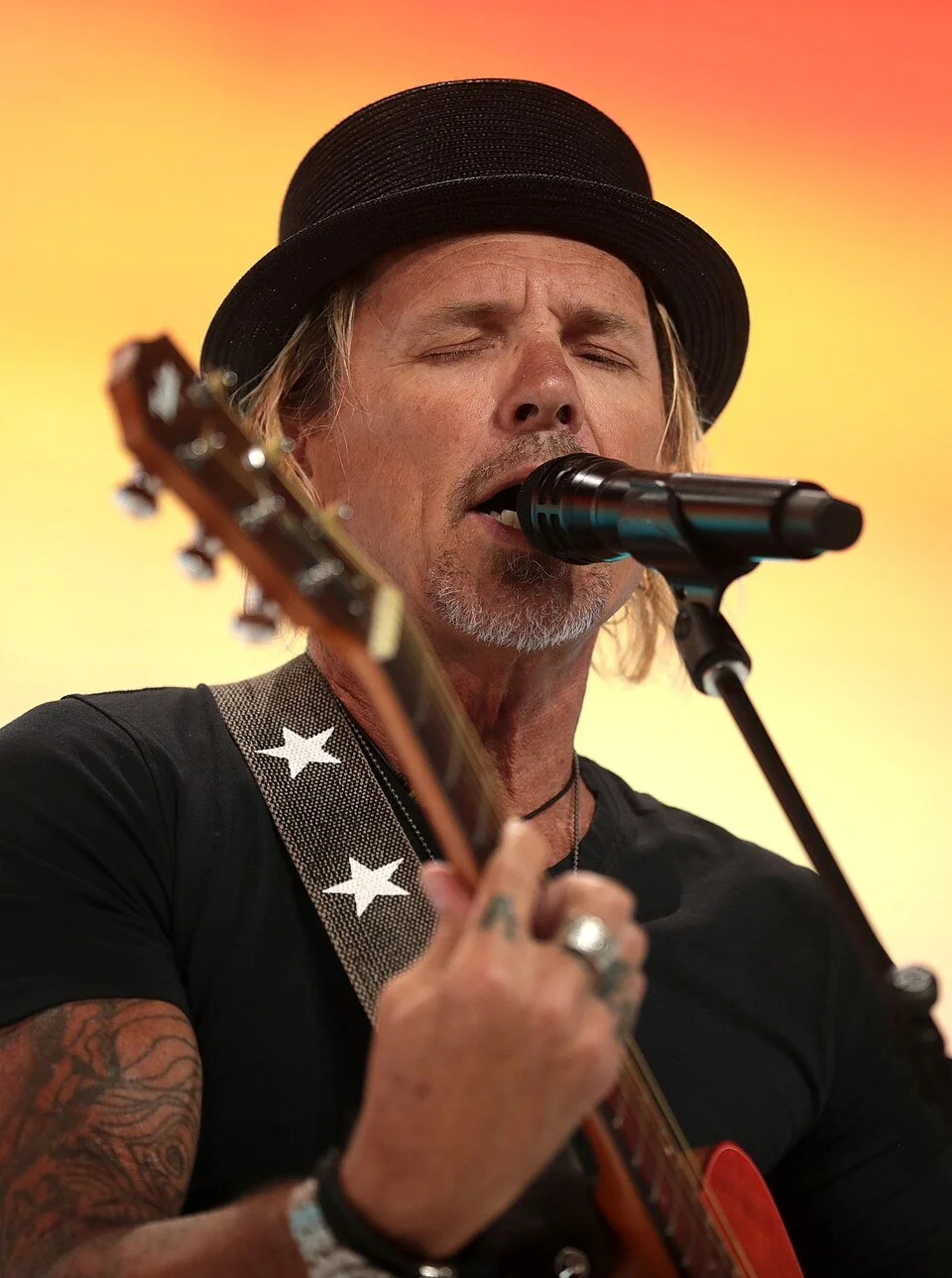 Male musician with closed eyes singing into a microphone while playing an acoustic guitar, wearing a black hat and black shirt, against a warm yellow-orange background.