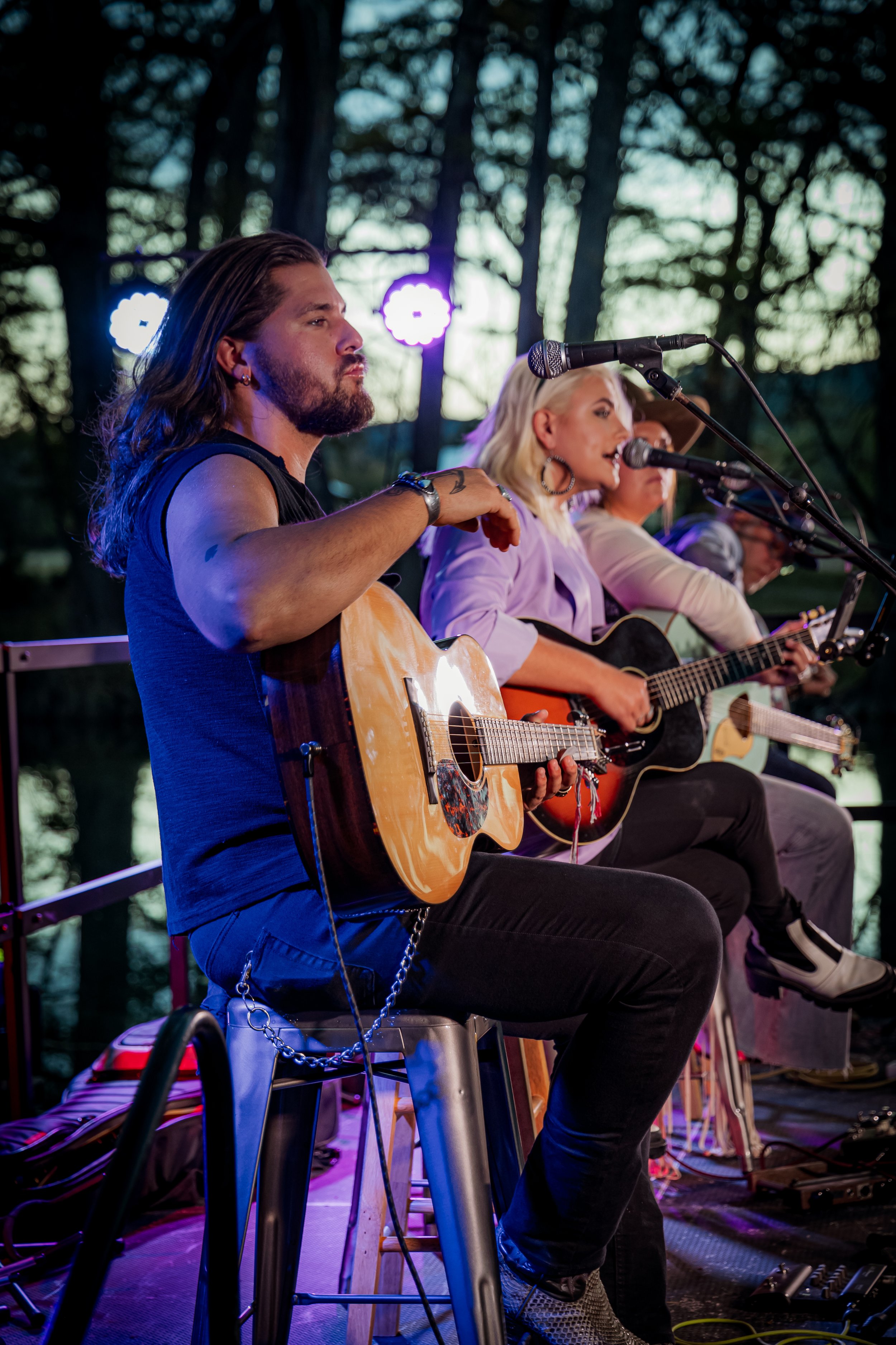 Three musicians sitting on stools playing guitars on an outdoor stage at sunset, with trees in the background and stage lights shining.