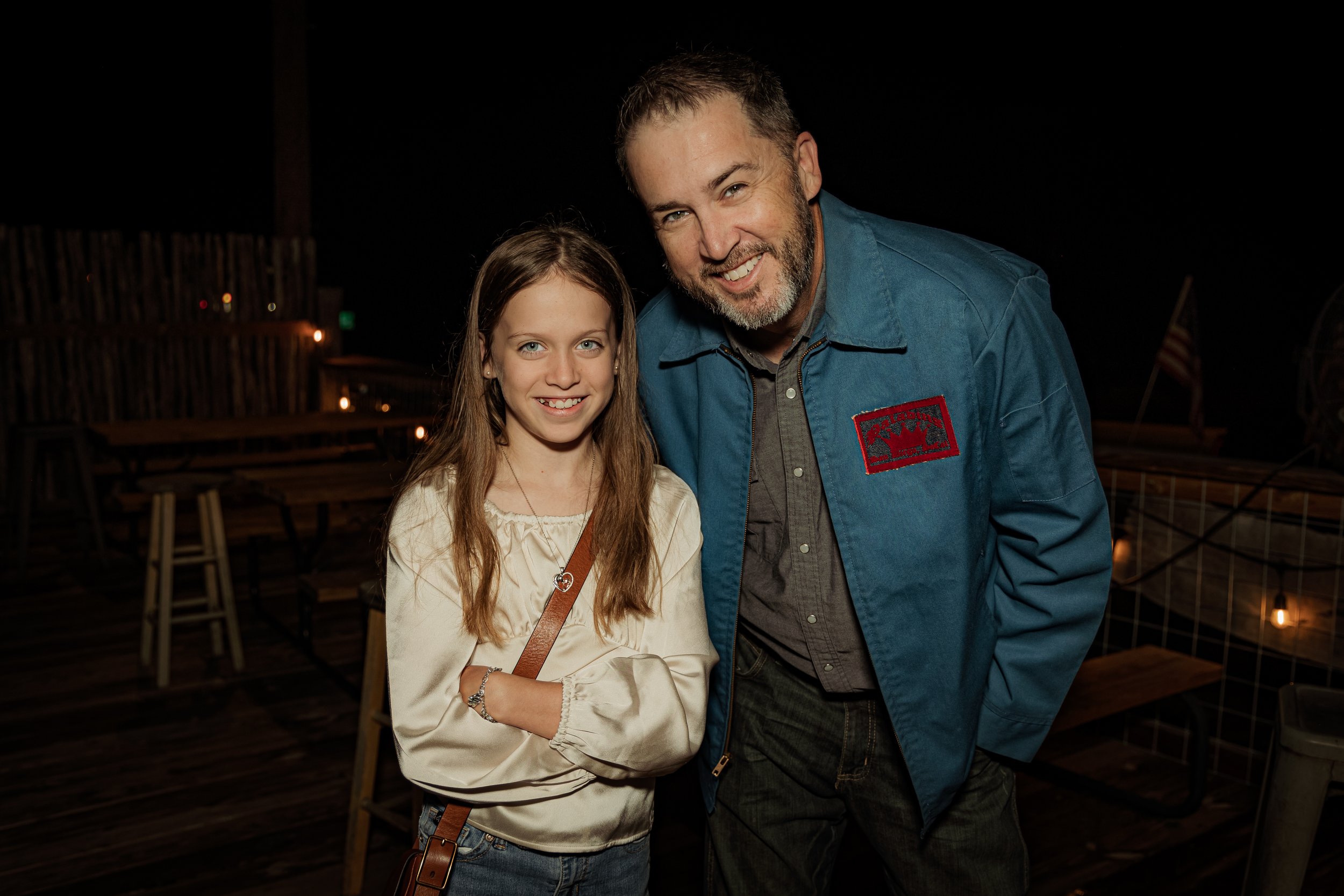 A man and a girl smiling at night on a wooden deck with string lights, with American flags in the background.