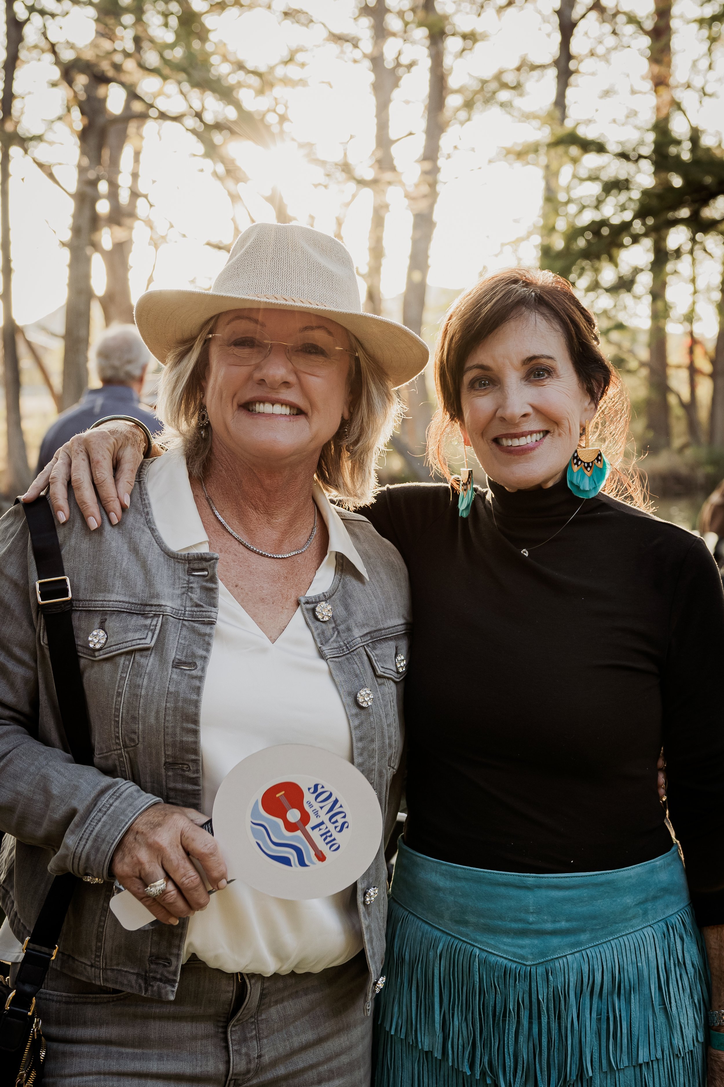 Two women smiling outdoors at sunset, one wearing a hat and holding a fan that says 'Songs of the Fry' with a lifeguard flag design, surrounded by trees.