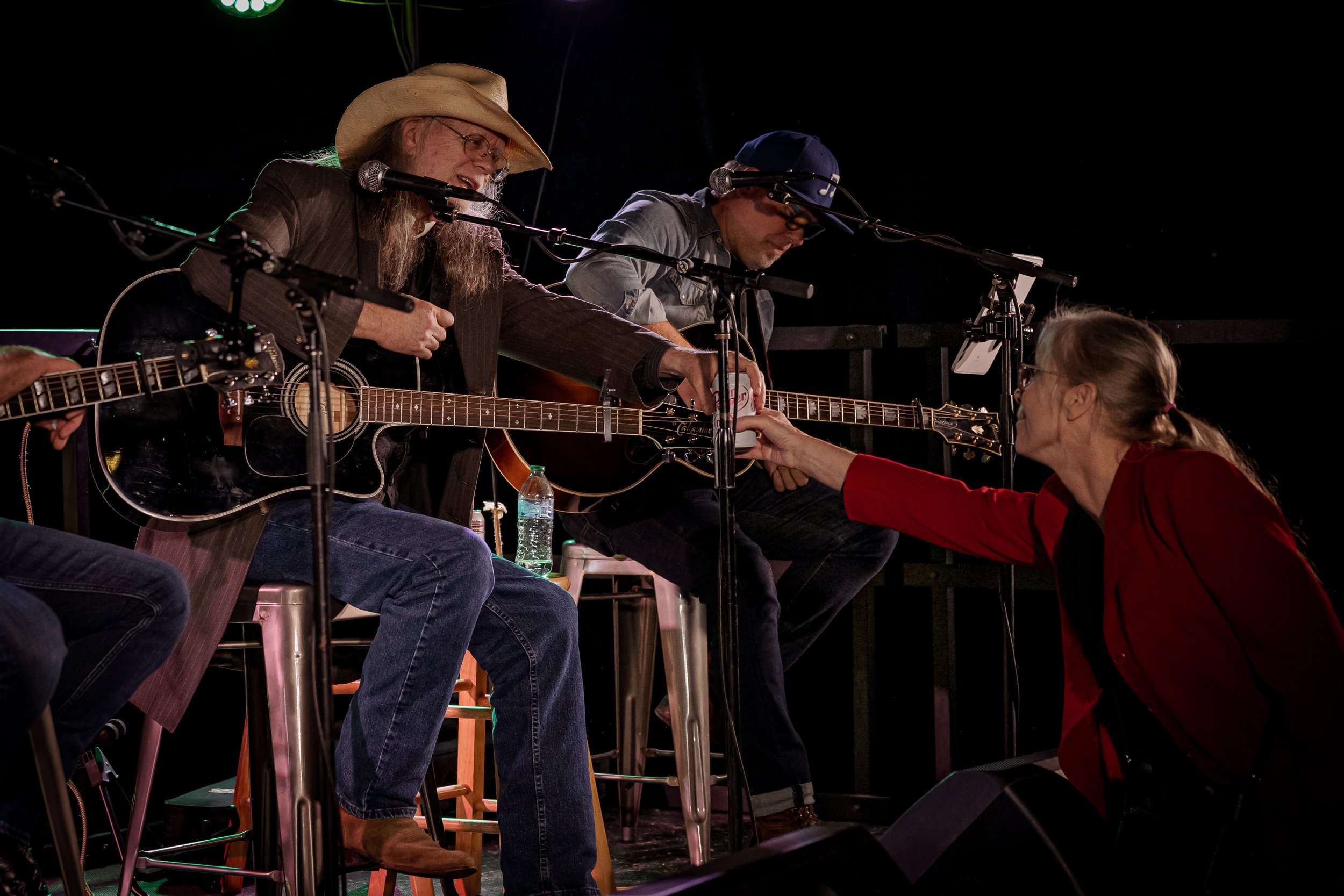 Woman in red jacket handing money to seated musicians with guitars during a performance.
