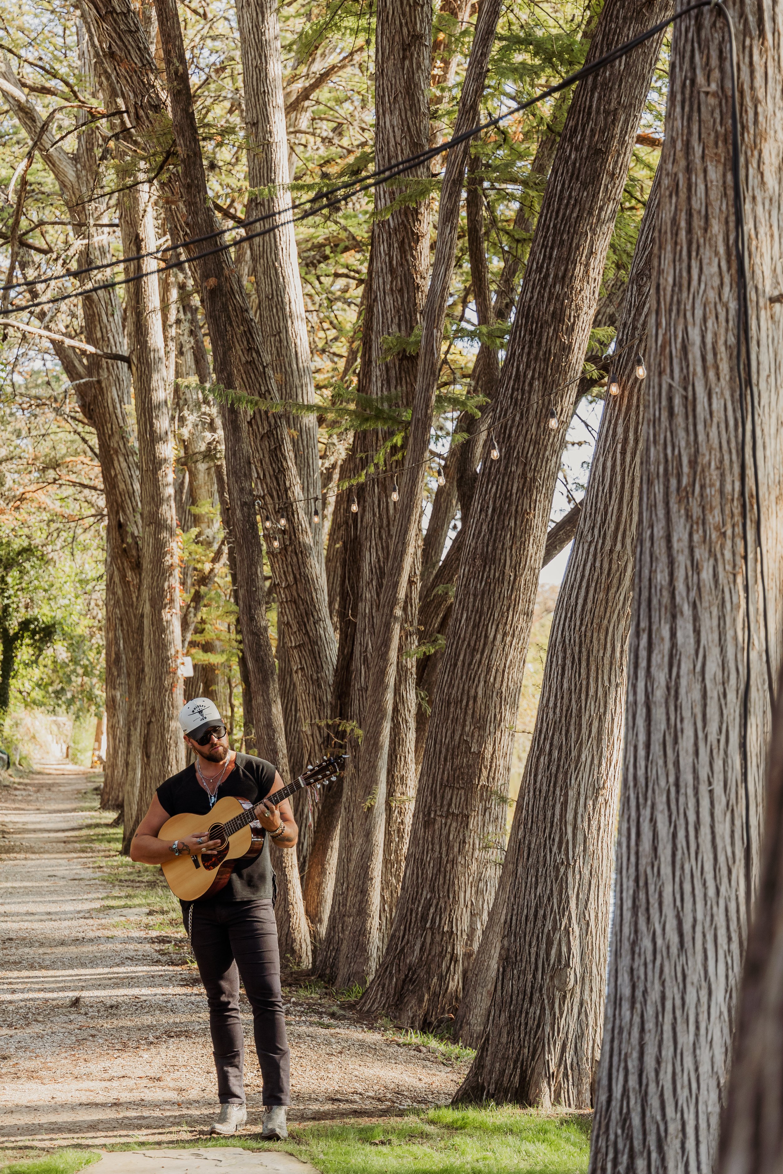 A man wearing a hat, sunglasses, and black clothing playing an acoustic guitar on a wooded path decorated with string lights.