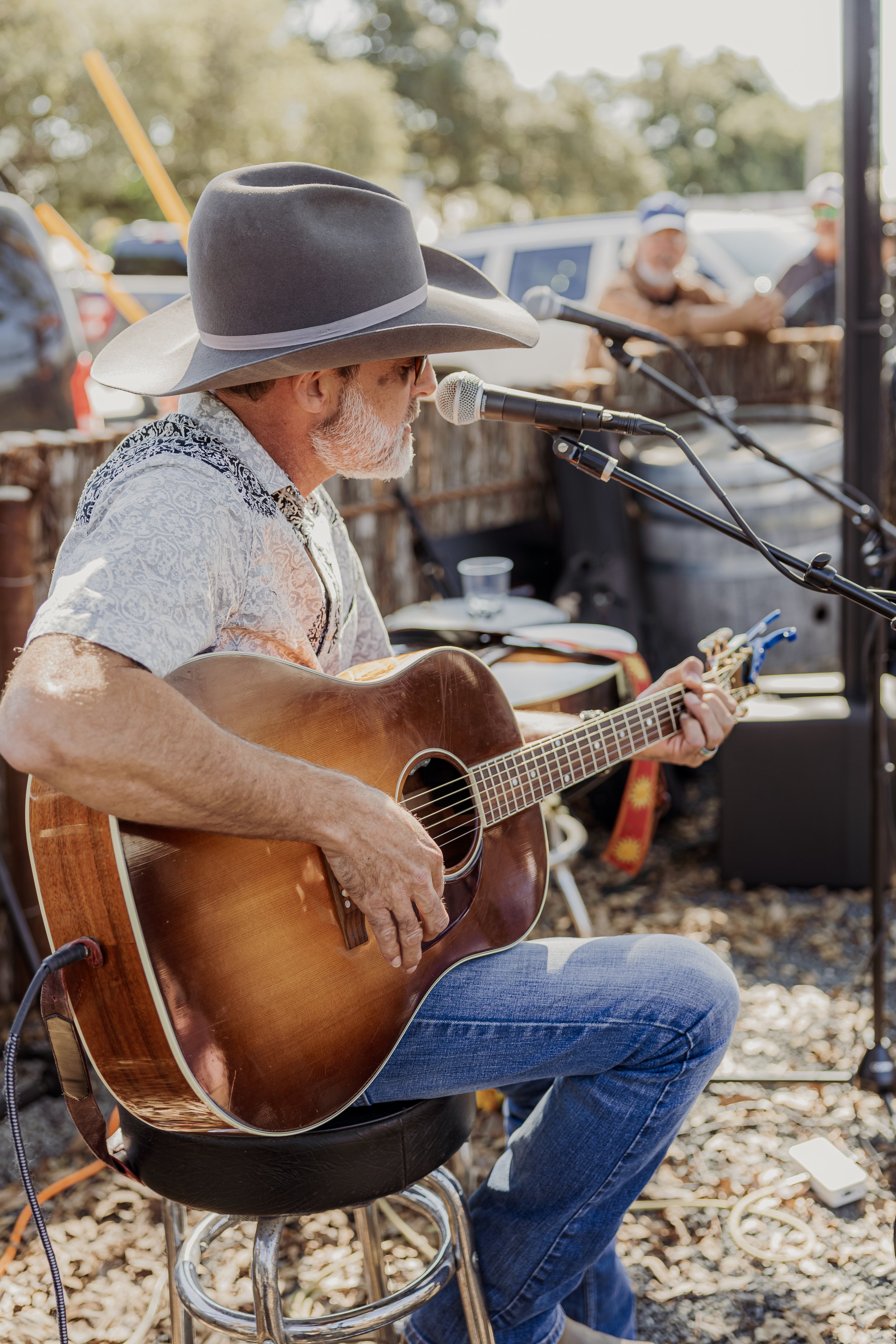 Man wearing a cowboy hat playing an acoustic guitar and singing into a microphone at an outdoor event.