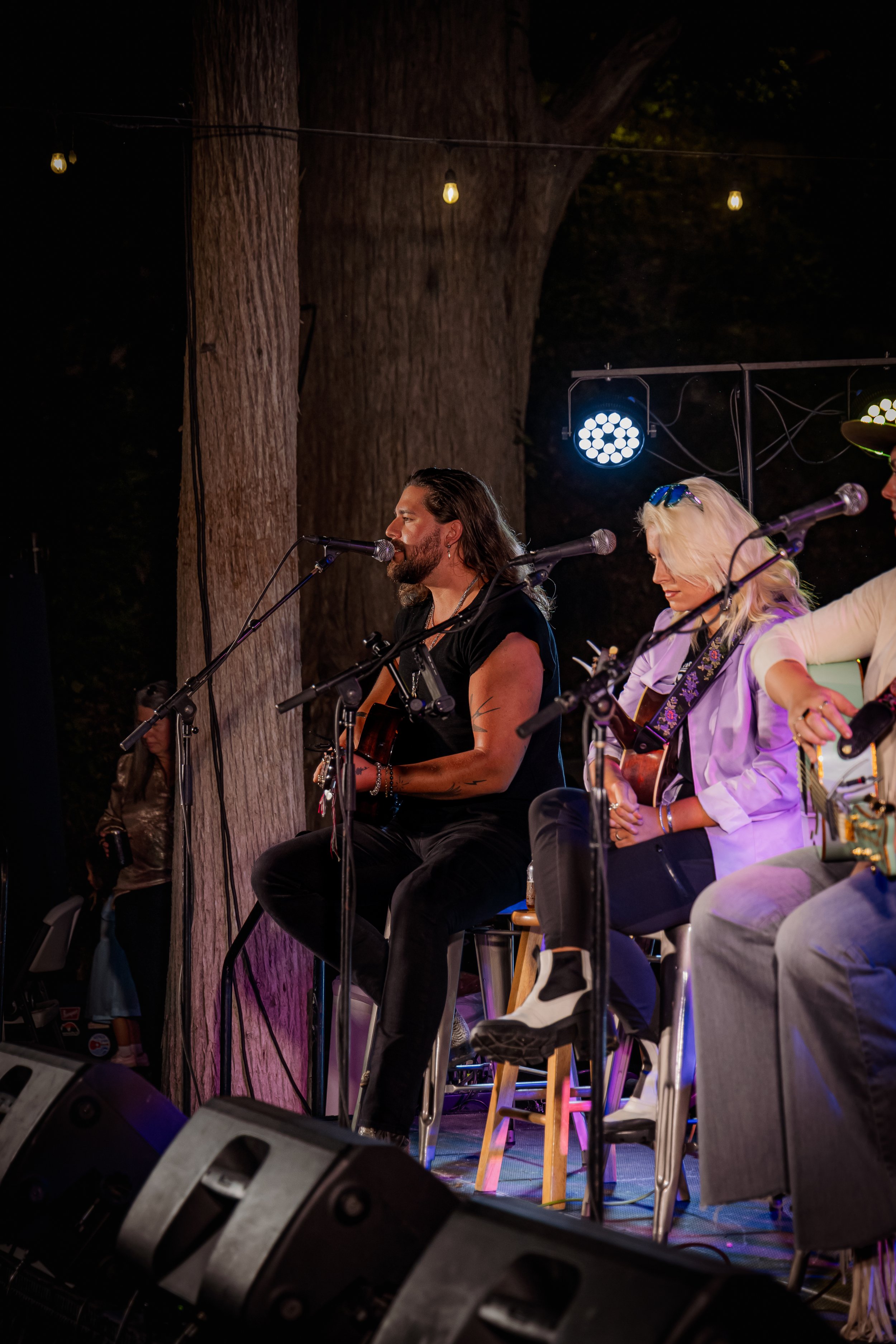Musicians performing on stage outdoors at night, seated with guitars and microphones, under string lights and large trees.