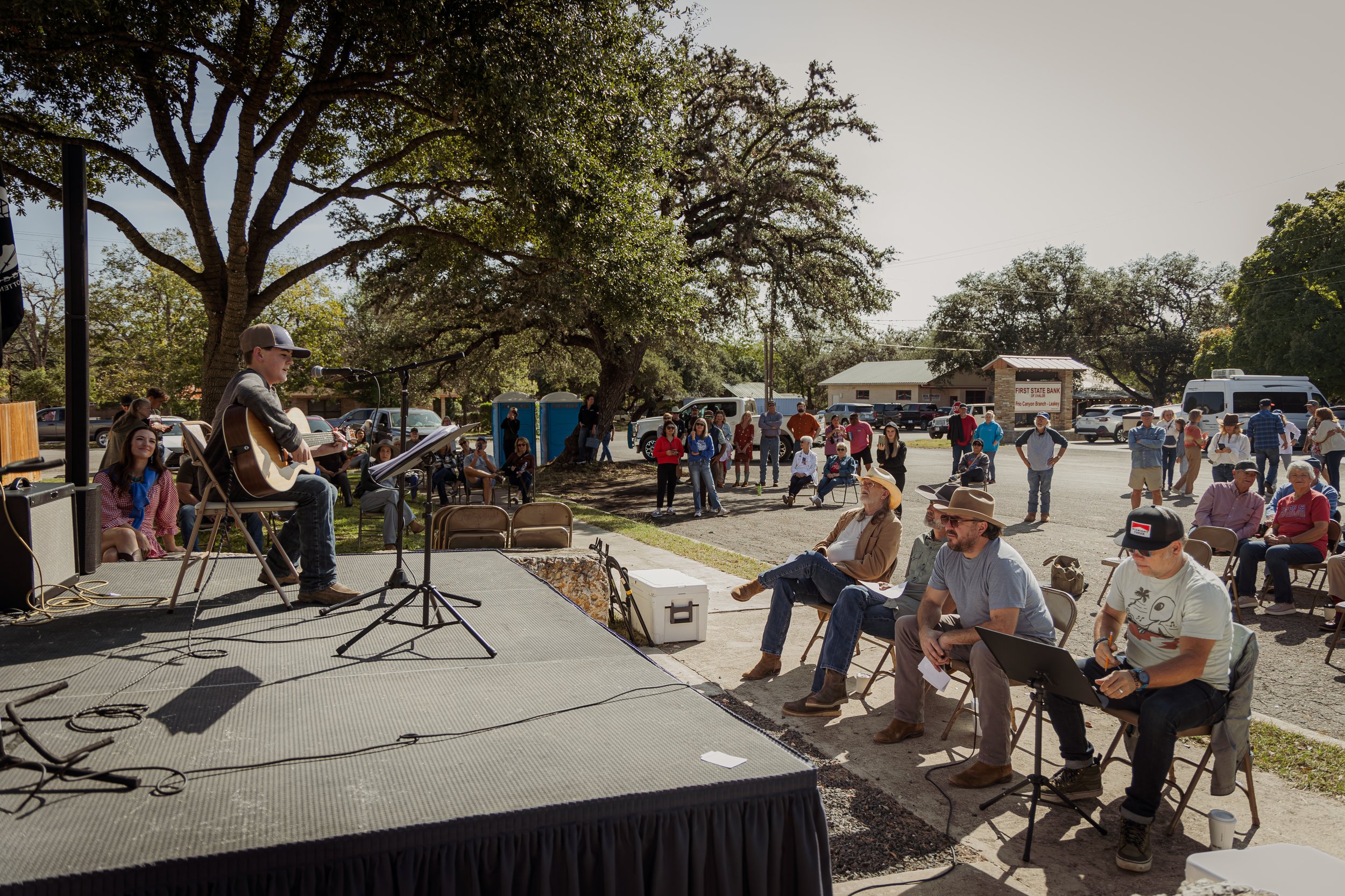 Young boy playing guitar and singing on stage at an outdoor event, with several seated and standing audience members watching under trees on a sunny day.