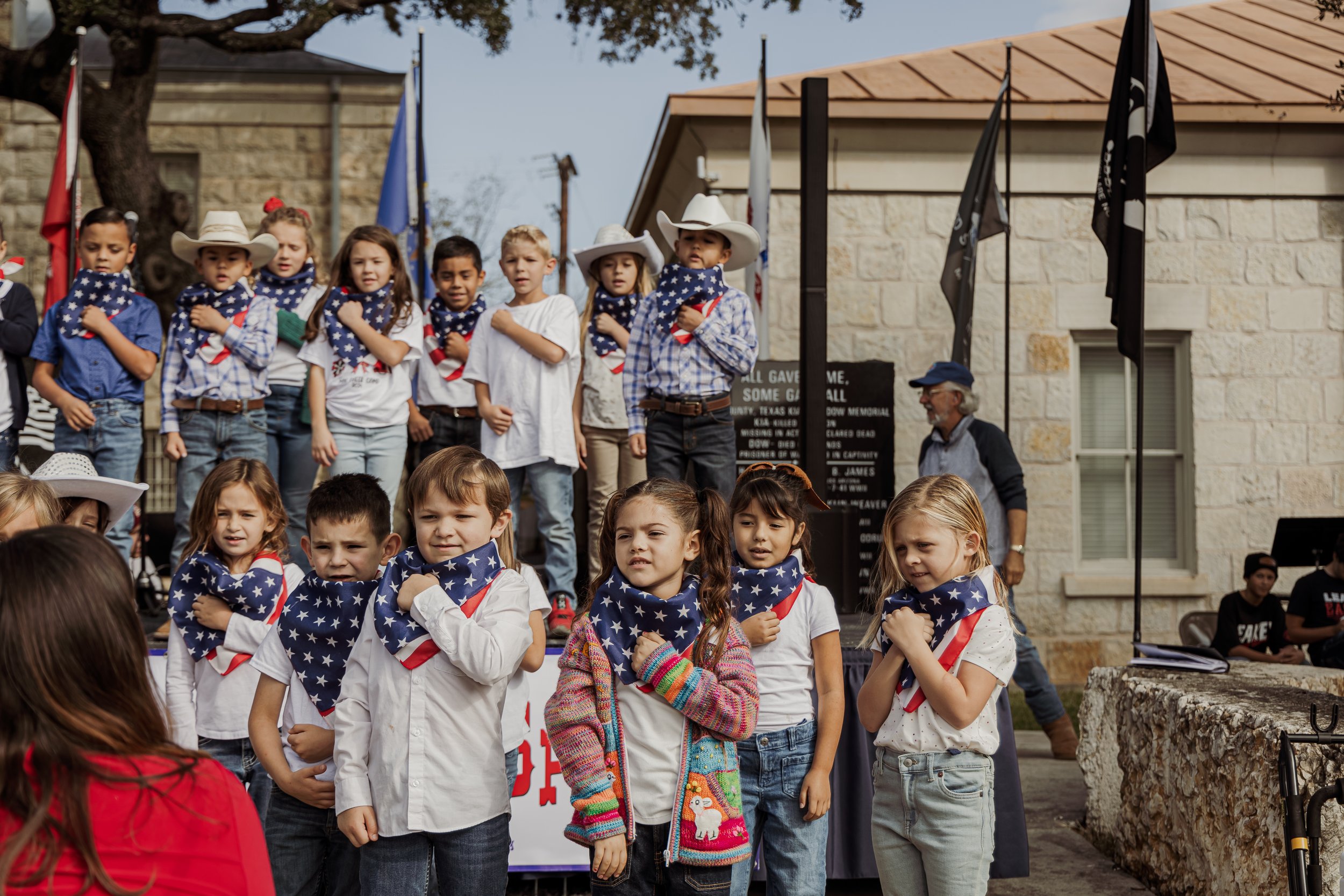 Children participating in a patriotic ceremony, wearing bandanas with American flag designs, some with cowboy hats, on a stage in front of a building with flags and a man observing the event.