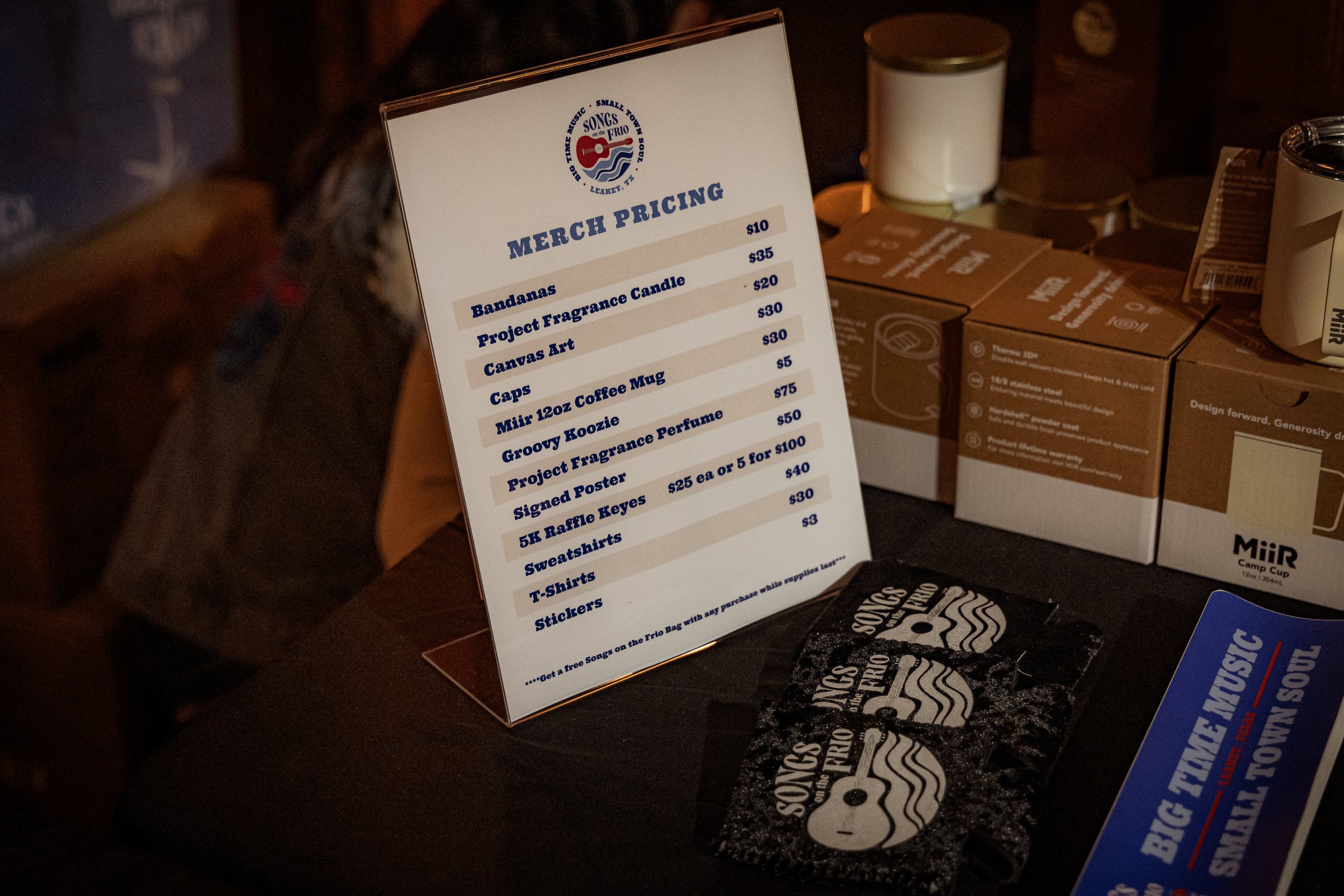 Merch pricing sign at Songs on the Frio music event listing various items with prices, including bandanas, candles, canvas art, caps, coffee mug, perfume, posters, keys, shirts, and stickers.