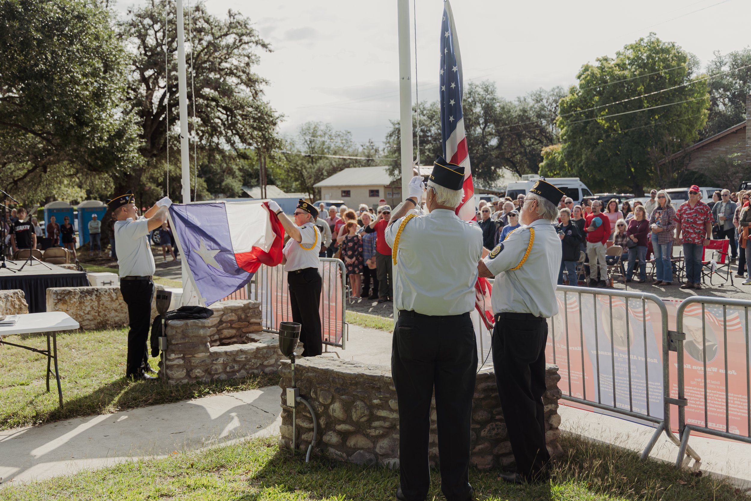 Four uniformed individuals are raising the Texas state flag and U.S. flag during a flag-raising ceremony at a community event, watched by a large crowd.