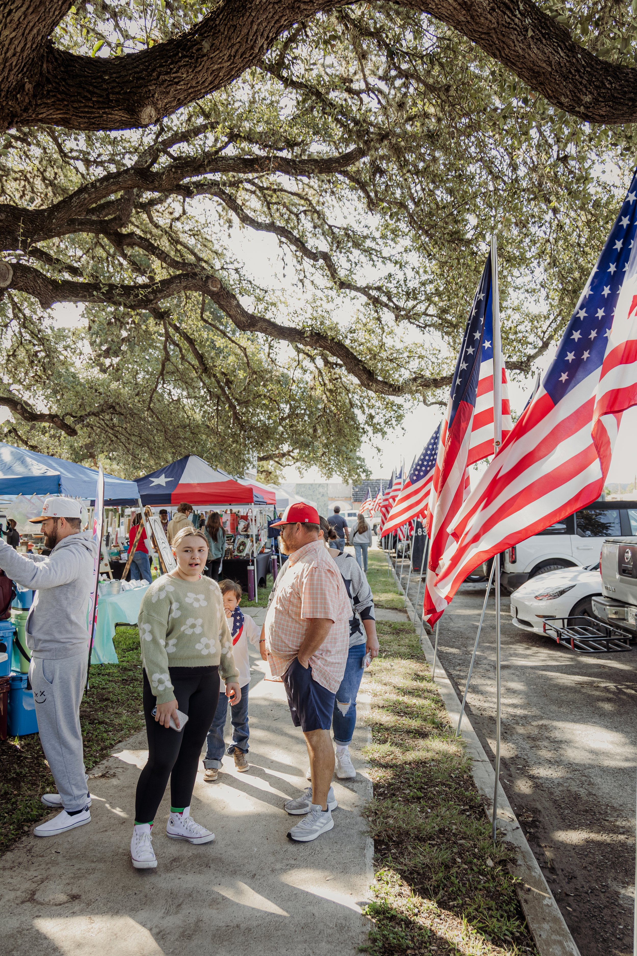 People at an outdoor event with American flags, tents, and vendors under large trees.