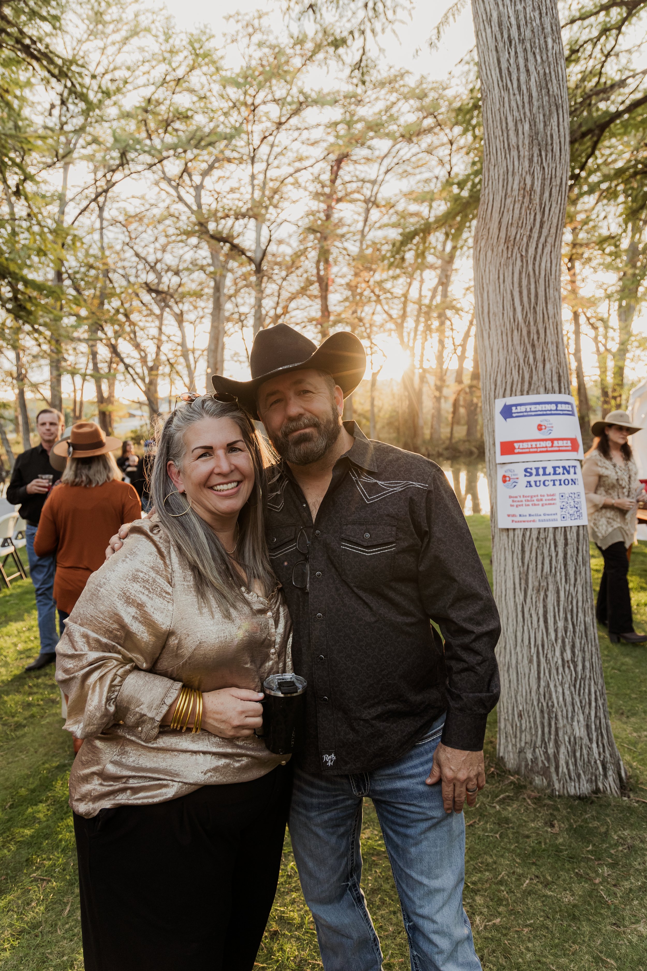 A smiling woman and man in cowboy attire standing outdoors during sunset, with other people in the background and signs on a tree nearby.
