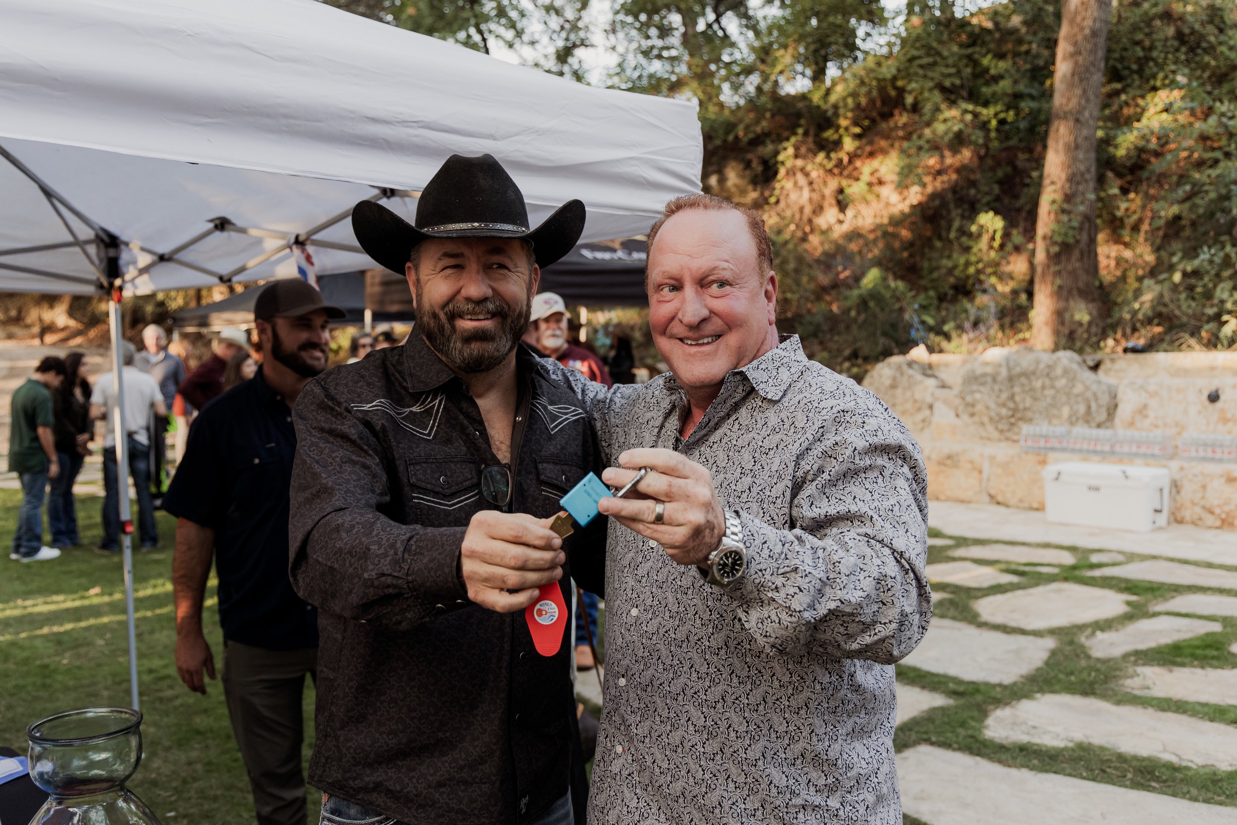Two men smiling and holding a lighter and a small blue container at an outdoor event, with a group of people and trees in the background.