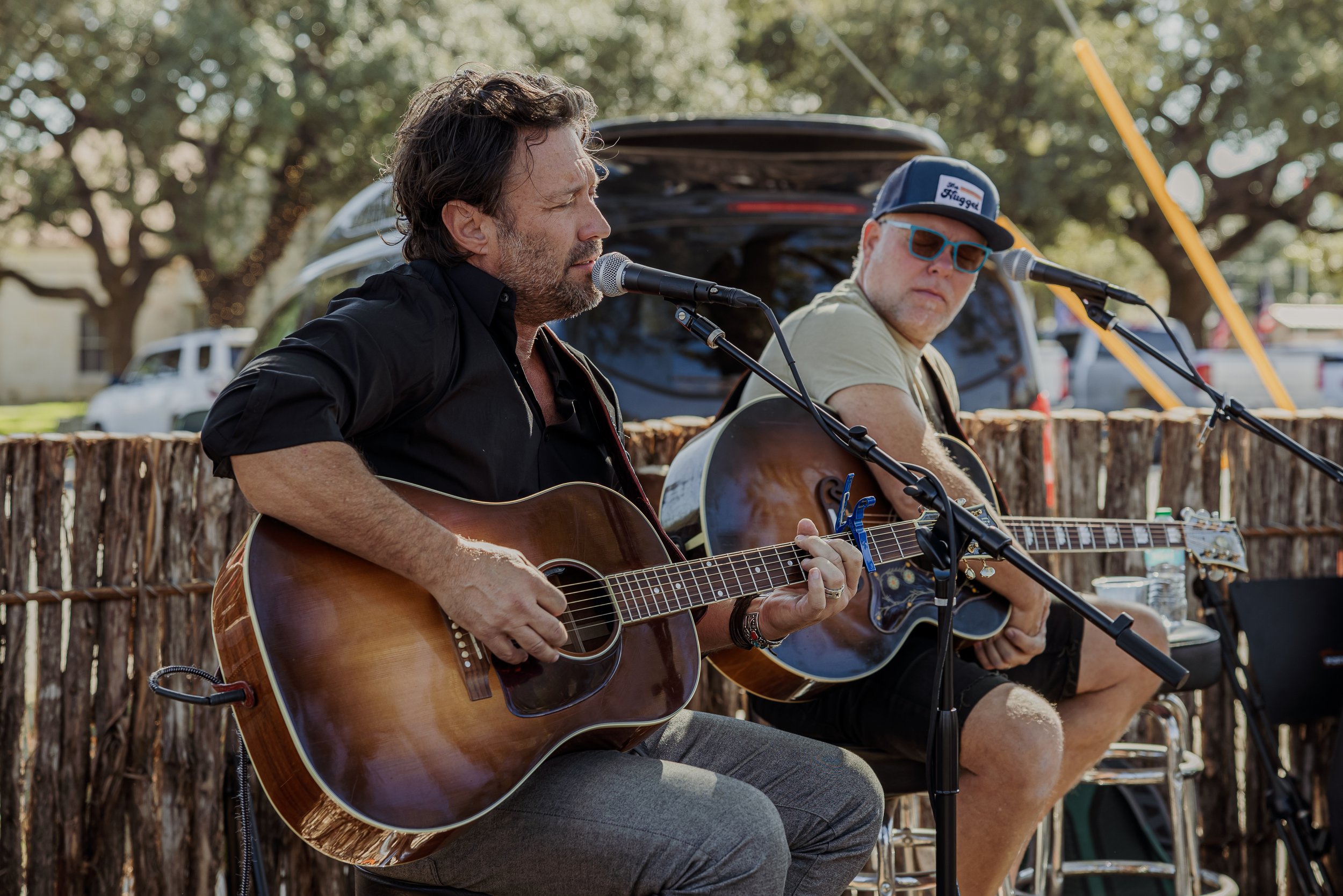 Two men playing acoustic guitars outdoors during a daytime performance, with a rustic wooden fence behind them and trees in the background.