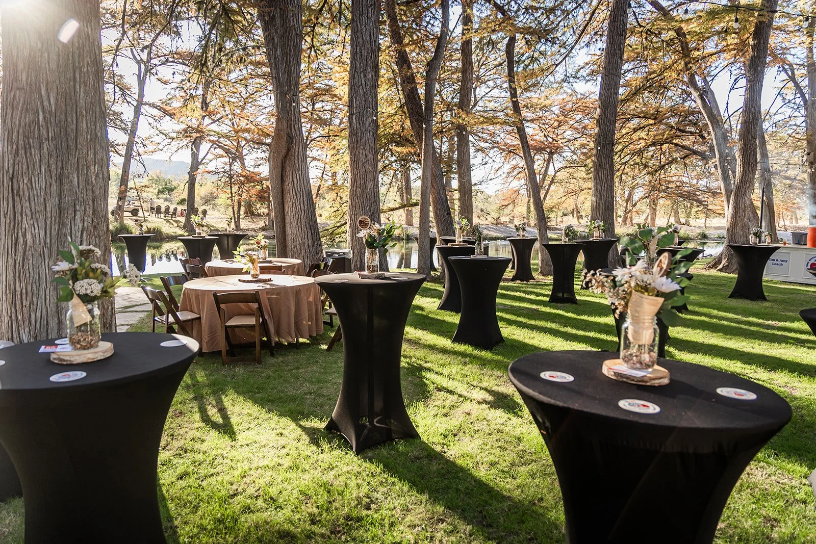 Outdoor event setup with tall cocktail tables draped in black and beige cloth, decorated with floral arrangements, situated on a grassy area among tall trees near a lake, sunlight filtering through the leaves.
