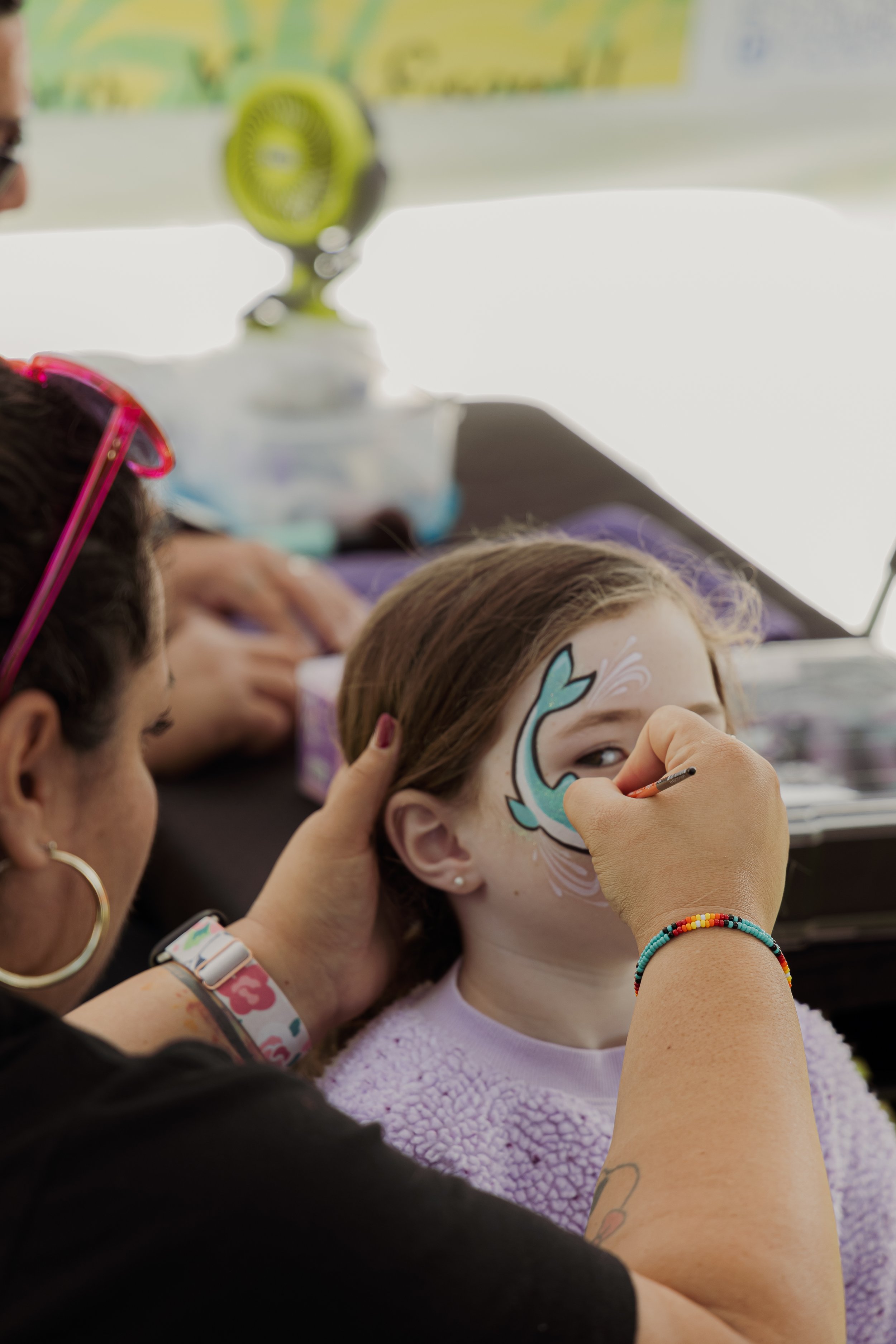 A young girl having her face painted with a blue and white whale design by an artist. The girl is wearing a purple fuzzy sweater, and the artist is wearing a black shirt and colorful bracelets.