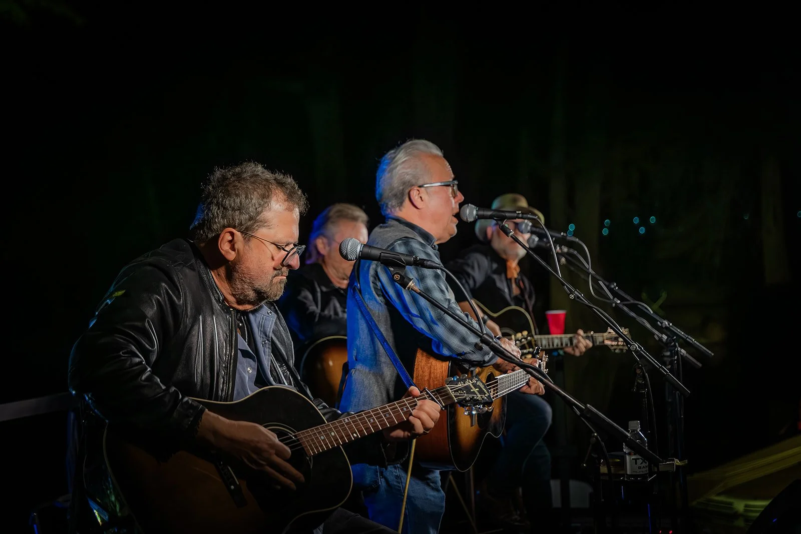 Four men sitting on a stage playing guitars and singing into microphones during a nighttime performance.