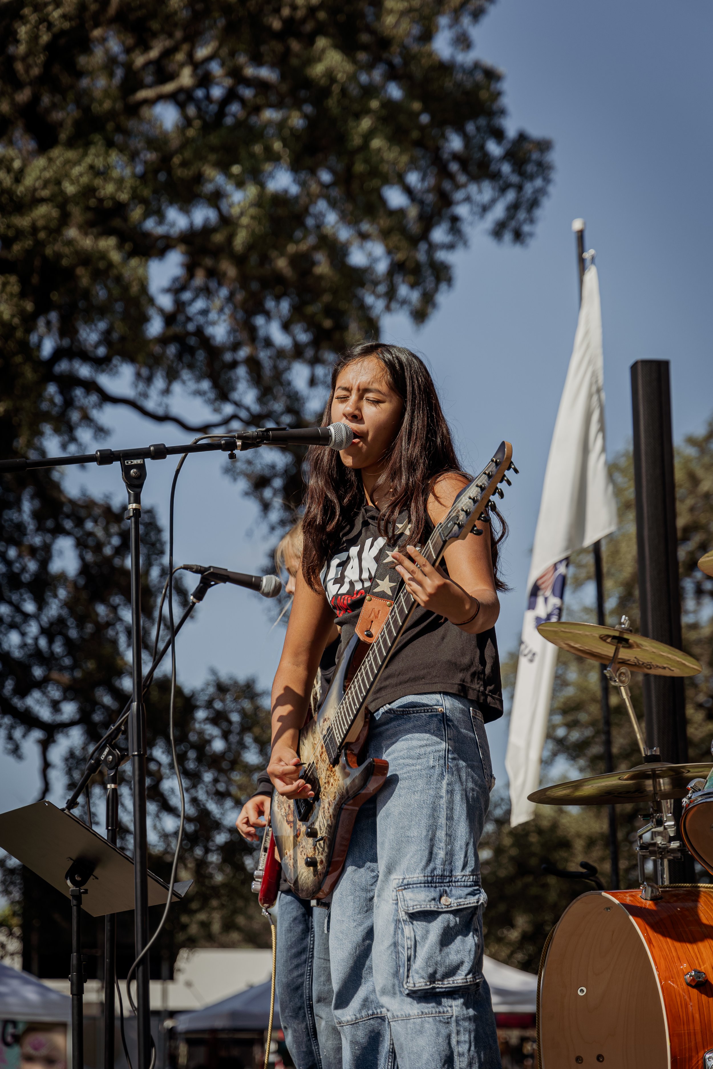 Young woman playing electric guitar and singing into microphone outdoors during a live performance.