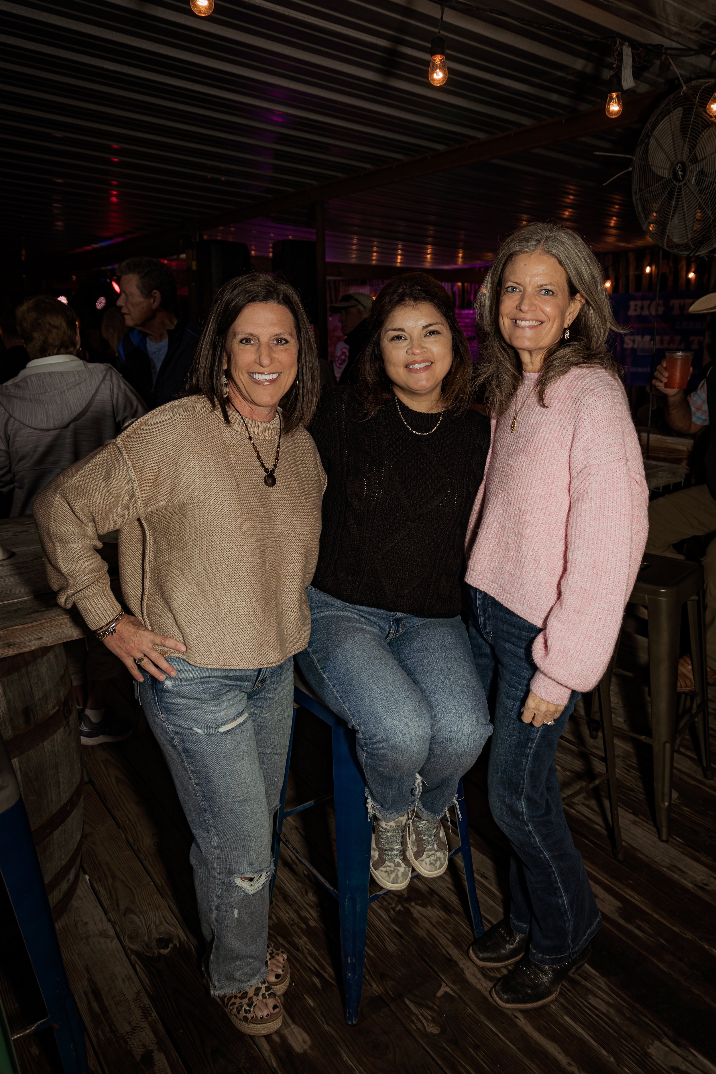 Three women smiling and posing together indoors, with dim lighting and other people in the background.