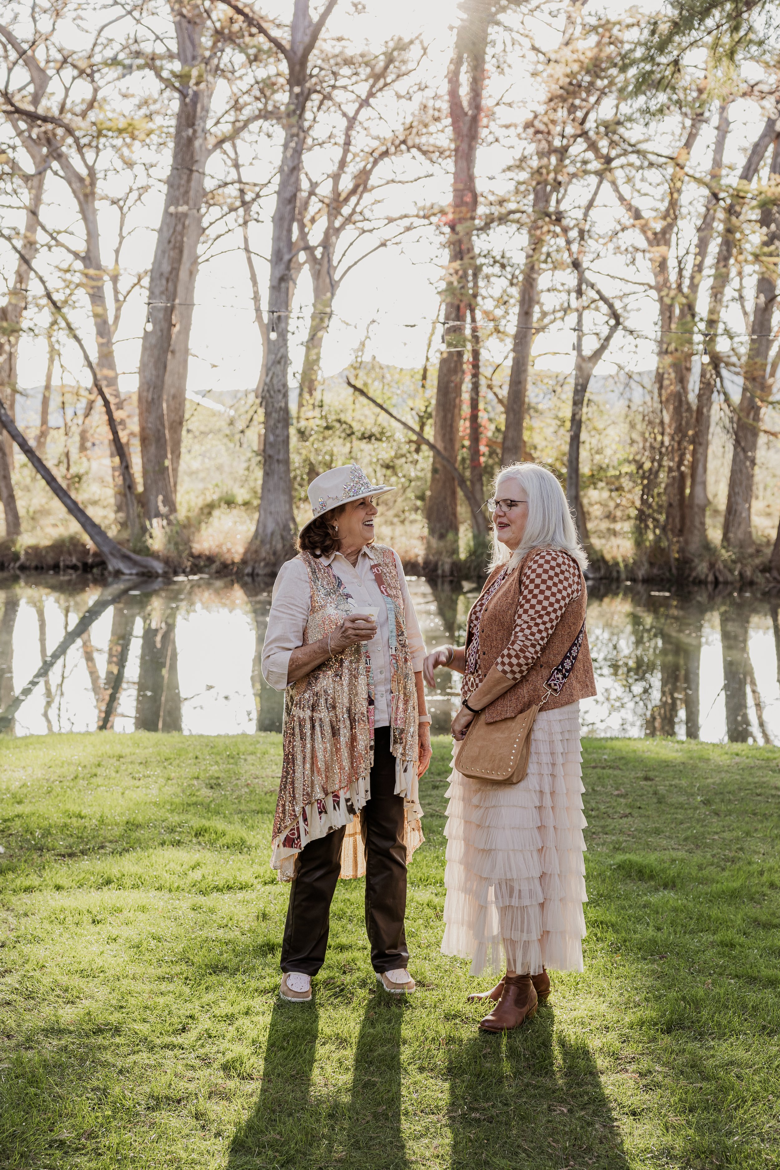 Two women are standing on grass near a river, engaged in conversation and smiling. They are dressed in casual, layered clothing with one wearing a hat. The background features trees with fall foliage and sunlight filtering through.