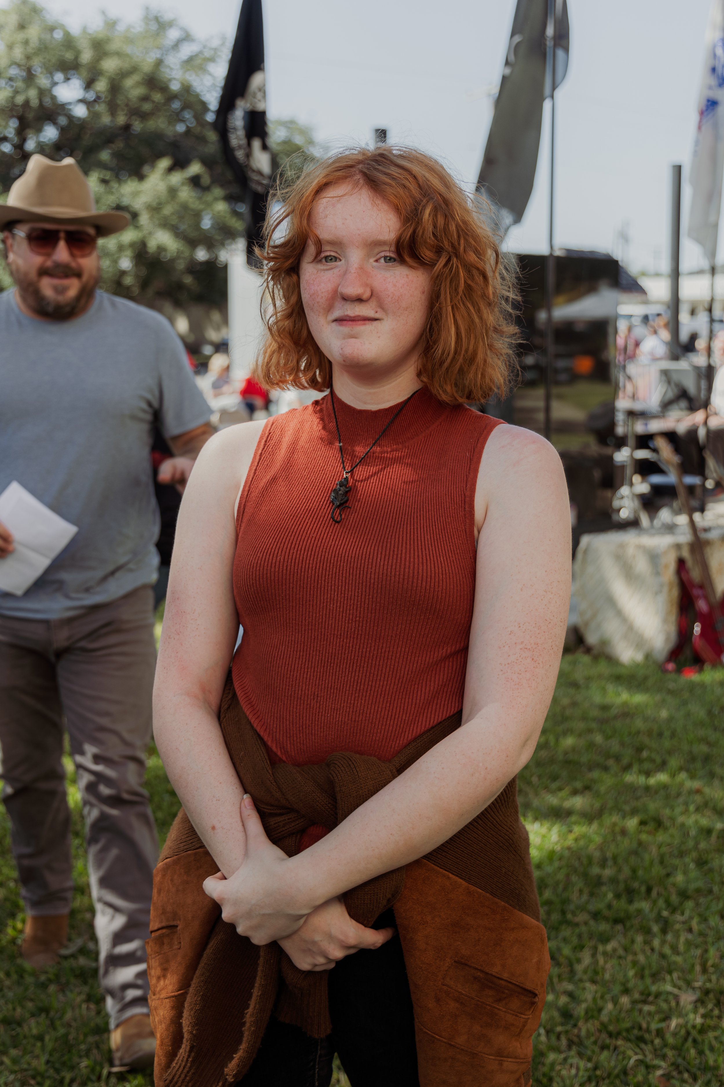 Young woman with red, curly hair wearing a sleeveless, rust-colored top and a brown sweater tied around her waist, standing outdoors at an event.